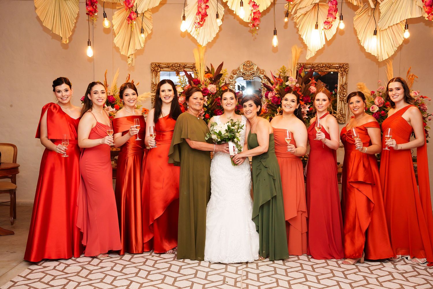 Bride in white gown poses with bridesmaids in red and olive green dresses, holding bouquets before floral backdrop.