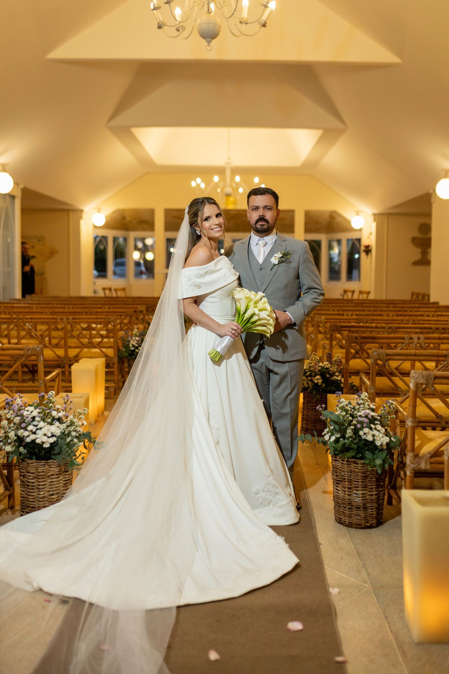 Bride and groom standing at the altar of an elegant chapel, bride in a flowing white gown with long veil holding calla lilies.