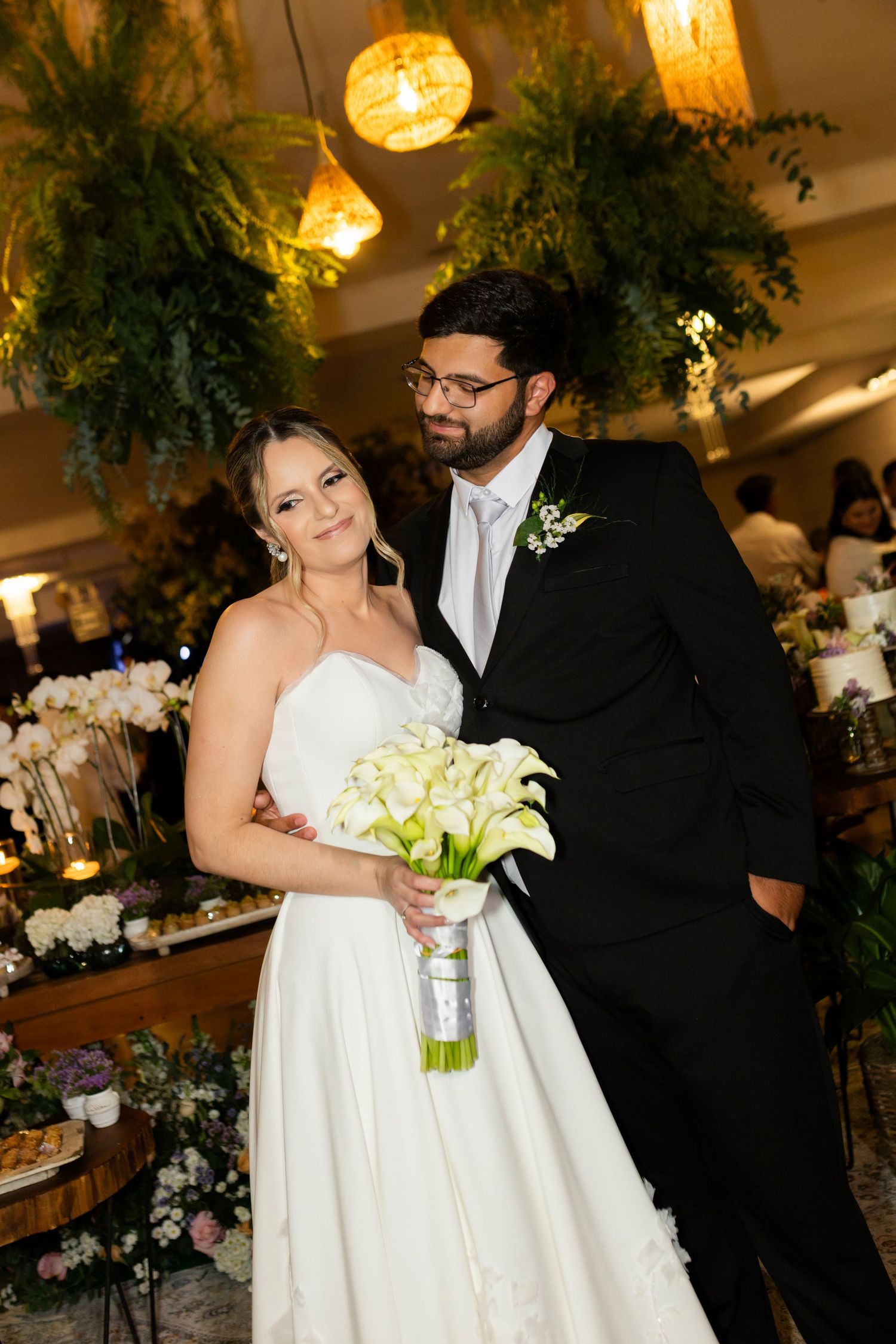 Bride in white gown holding calla lily bouquet stands with groom in black suit at elegant floral wedding reception.