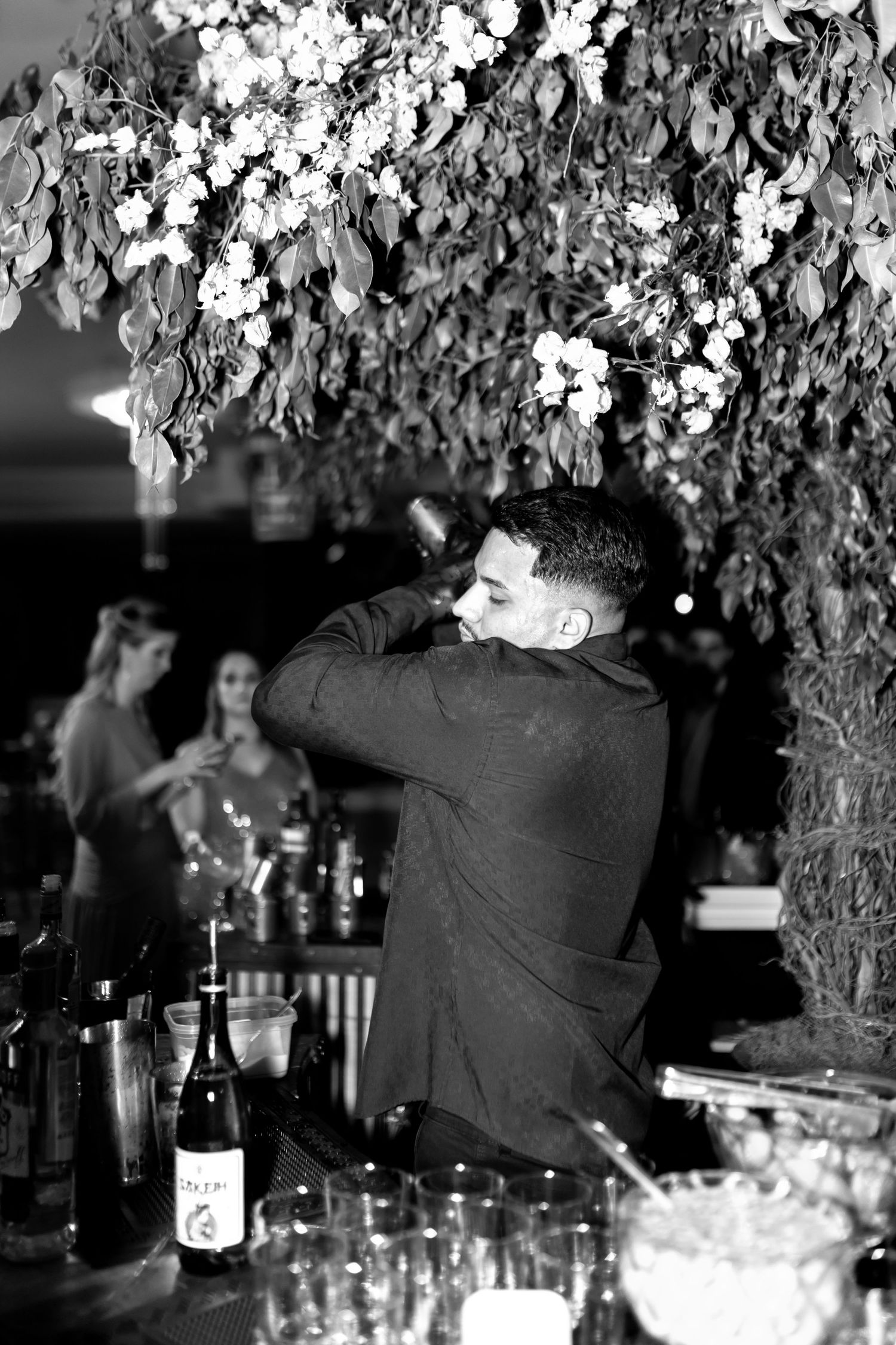 Bartender shaking cocktail under floral canopy at elegant event, black and white photography.