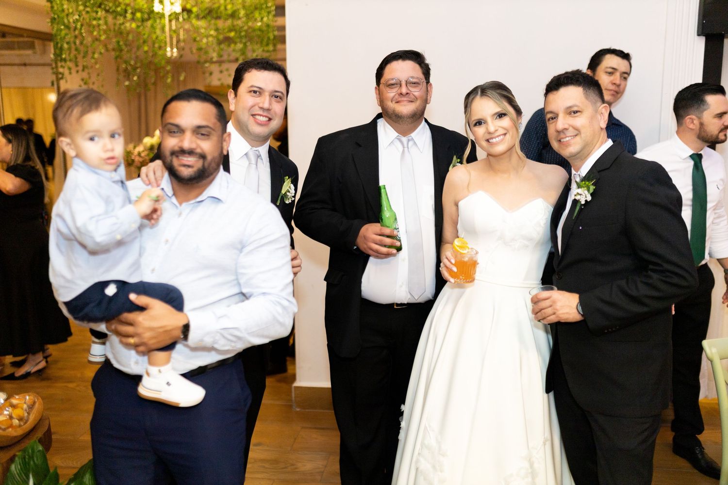 Wedding guests and newlyweds pose together smiling, bride in white gown holding drinks at elegant indoor reception.