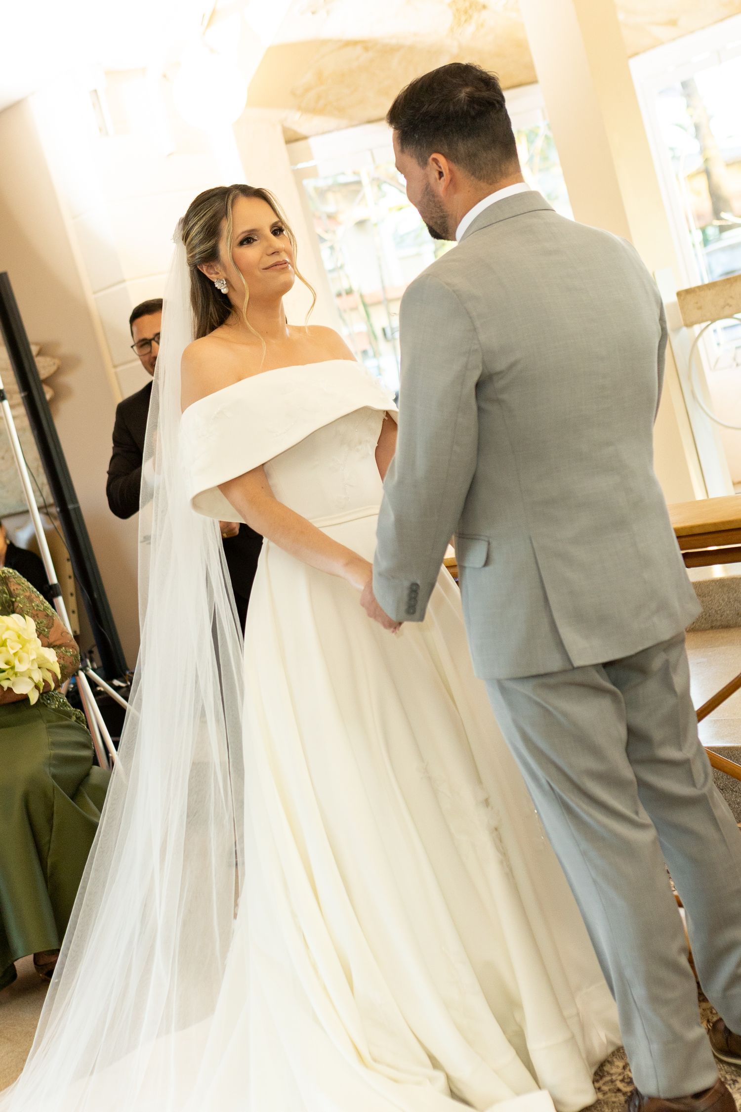 Bride in off-shoulder white gown with veil holds hands with groom in gray suit during wedding ceremony.