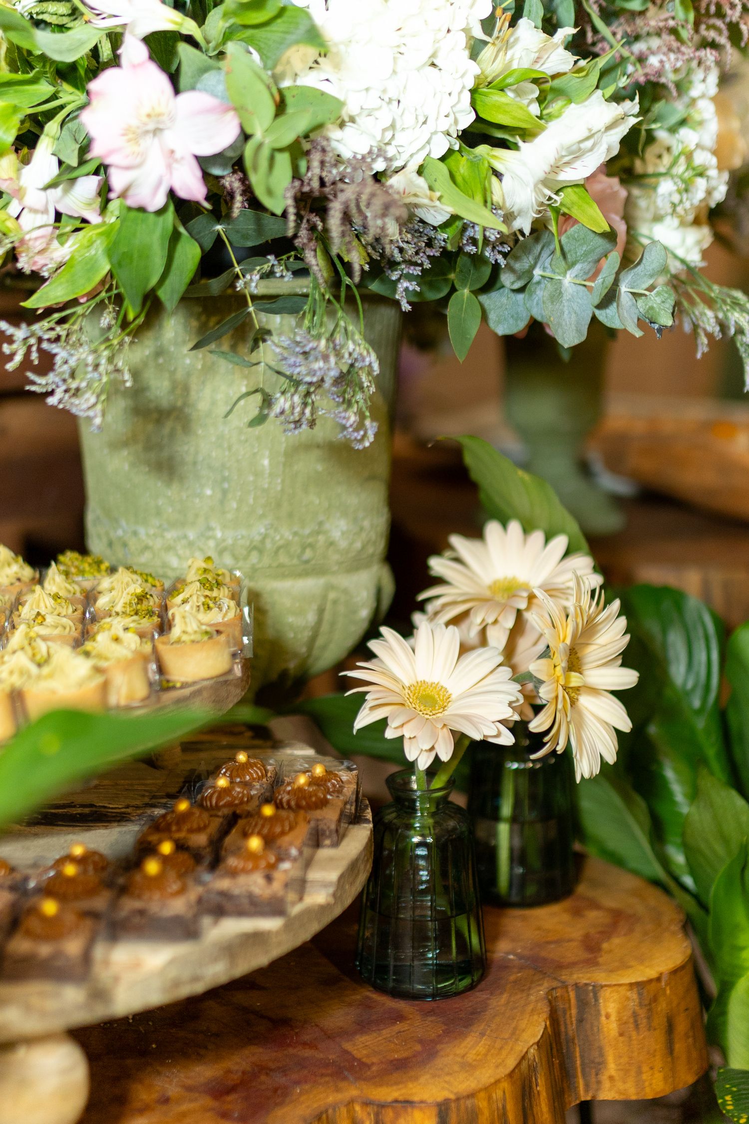 Elegant dessert table with gerbera daisies, cupcakes, brownies, and a lush floral arrangement in a green ceramic vase.