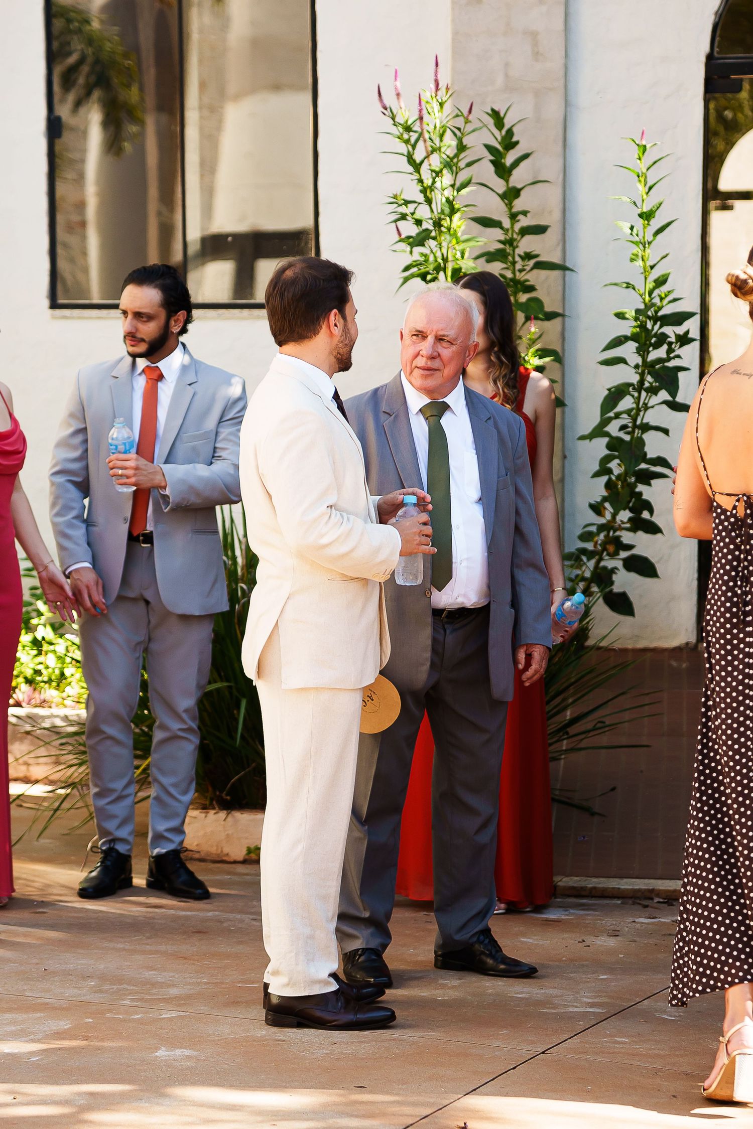 Elegantly dressed guests mingle outdoors at a wedding reception, with men in suits conversing near lush greenery.