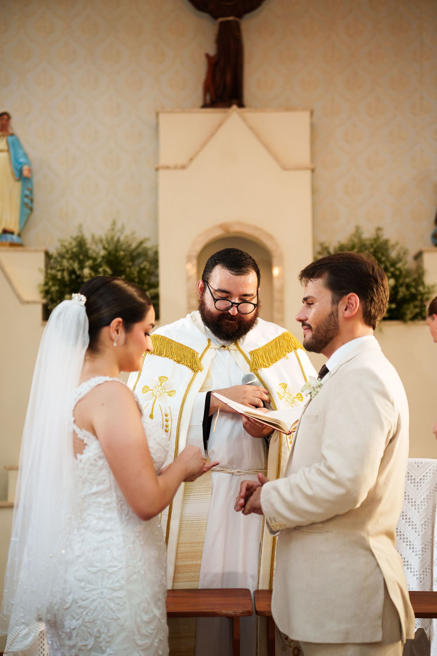 Bride and groom exchanging rings during a Catholic wedding ceremony with a bearded priest reading from a book.