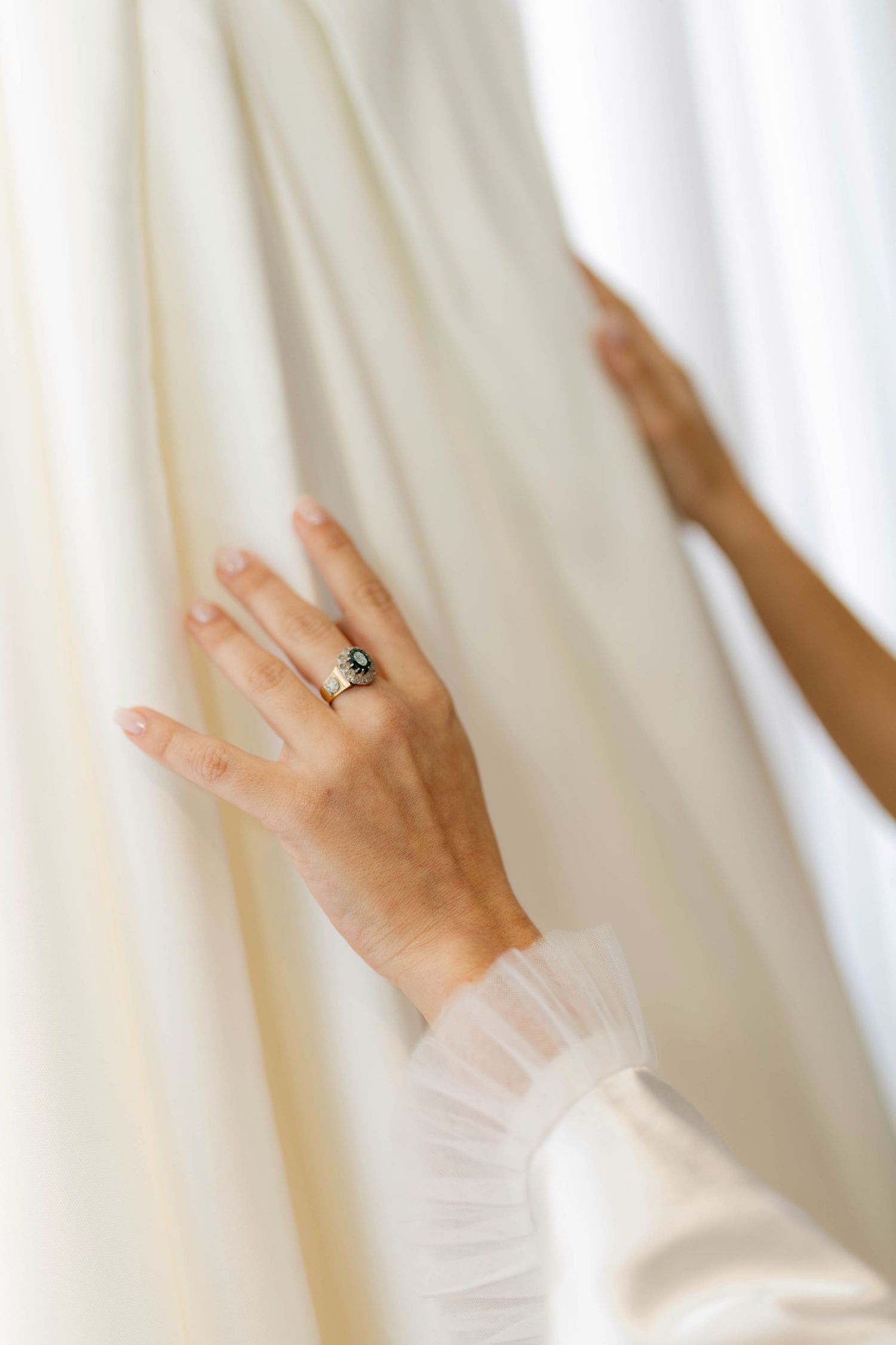 Close-up of a bride's hand wearing an elegant green gemstone ring, touching a white wedding dress.