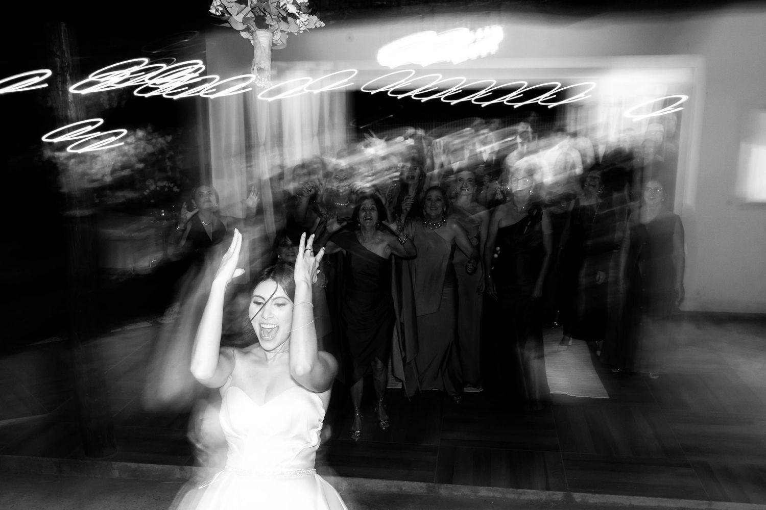 Black and white photo of a bride tossing her bouquet at a wedding reception with guests in the background.