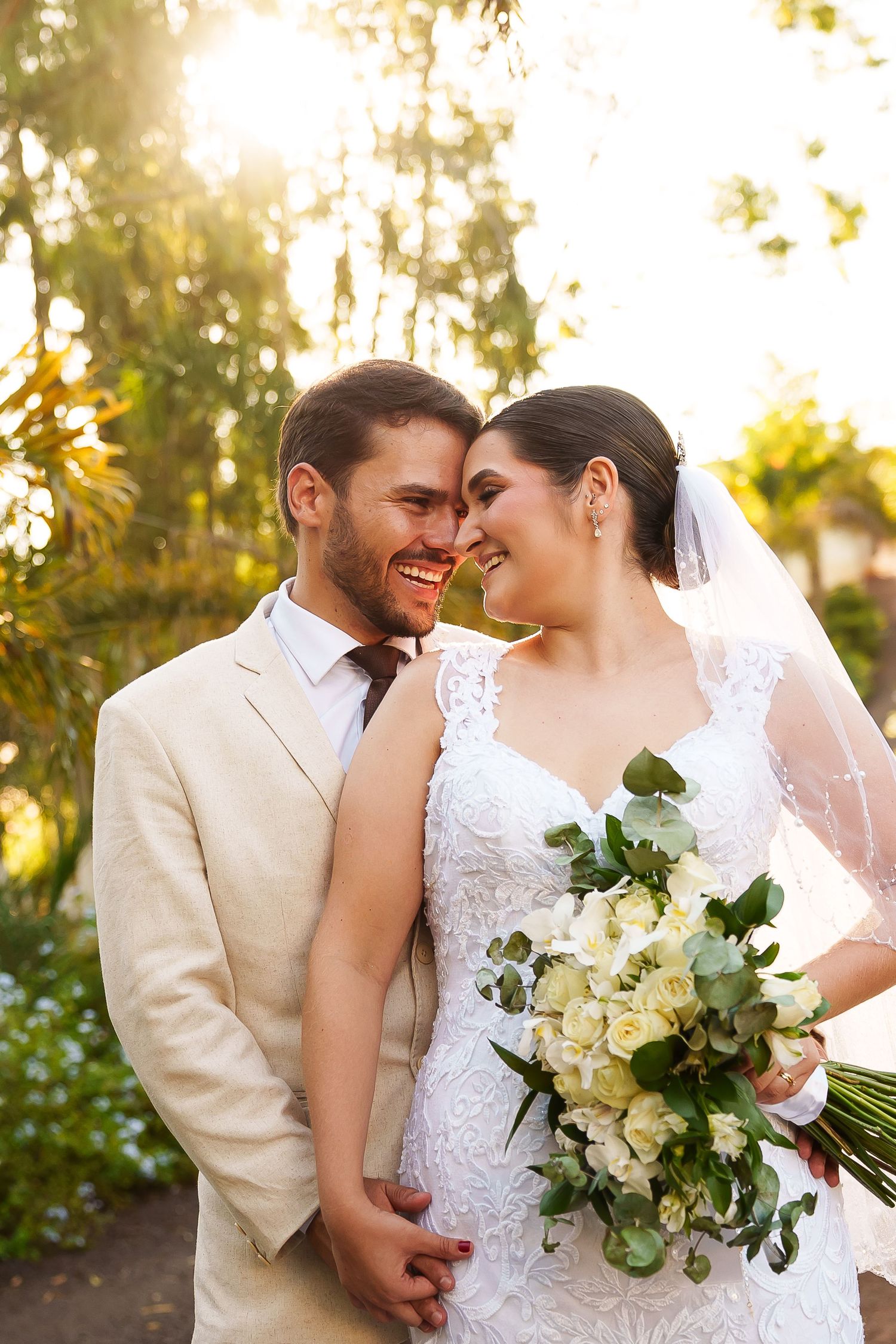 Smiling bride and groom touching foreheads in sunny garden, bride holding white rose bouquet, groom in beige suit.