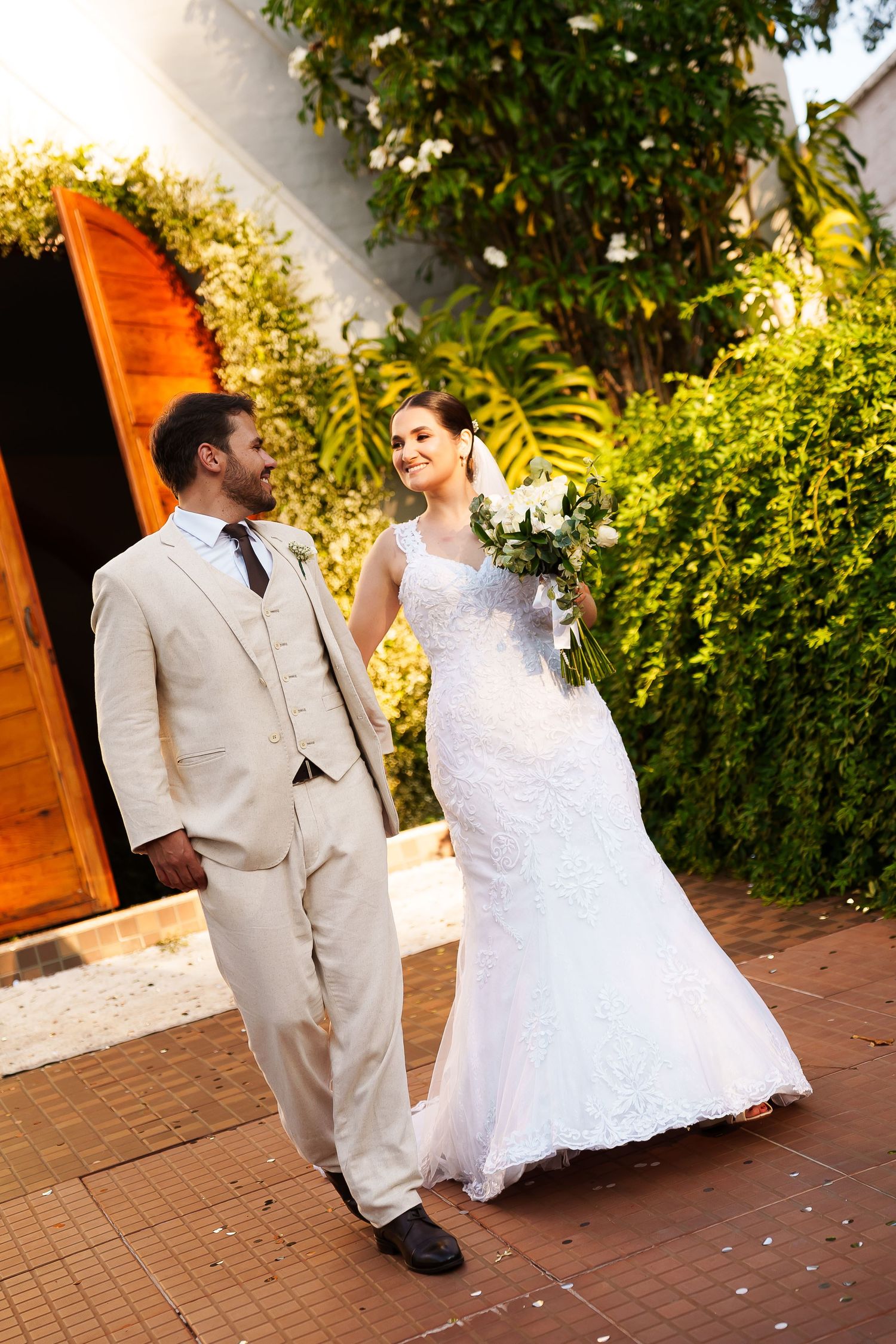 Bride in white lace gown and groom in beige suit walk together outdoors, smiling, with lush greenery behind them.