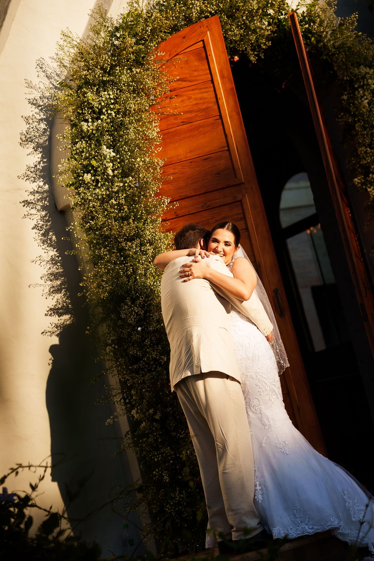 Bride and groom embrace in golden light outside a wooden door surrounded by lush greenery on their wedding day.