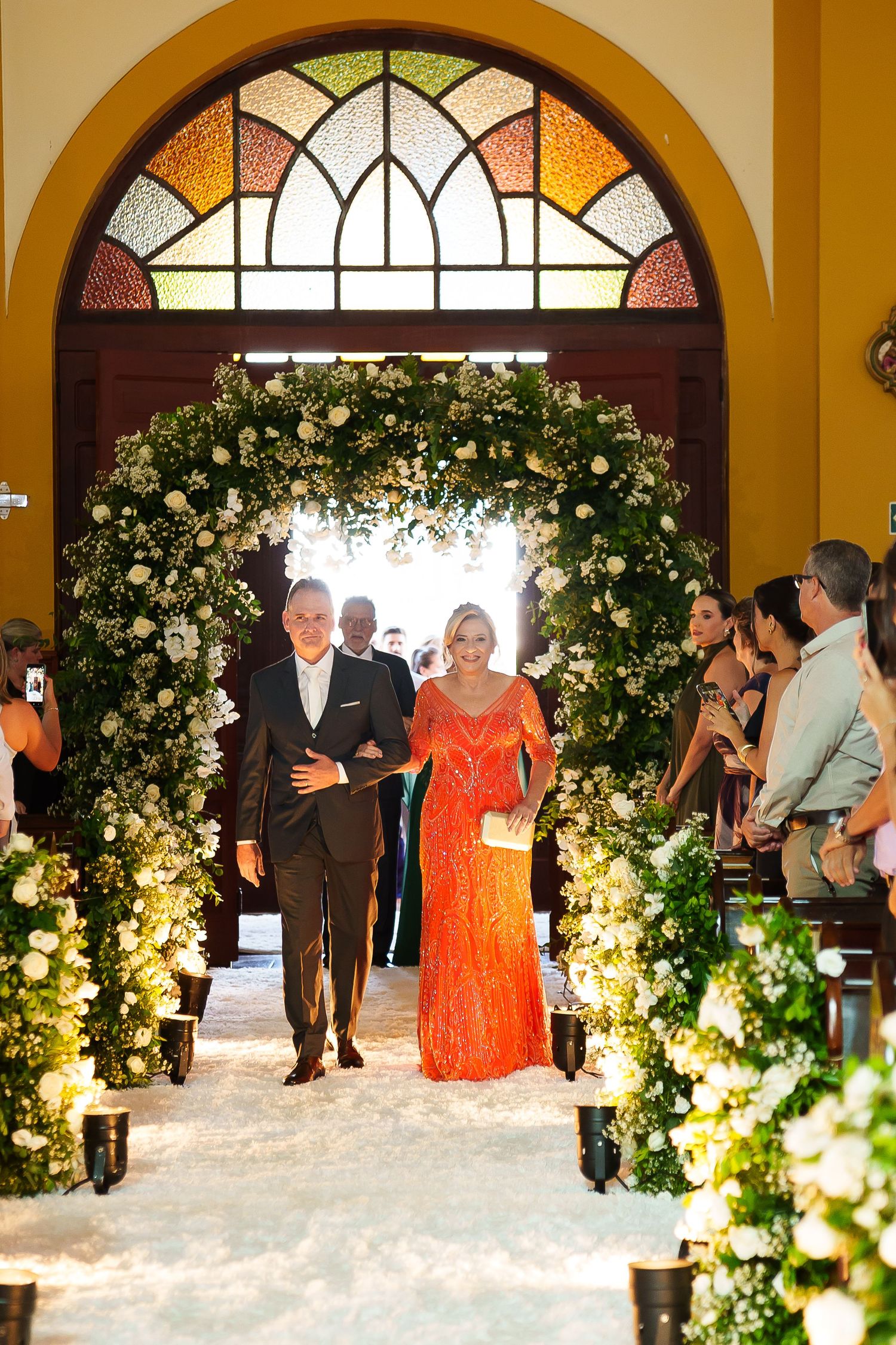 Elegant couple walks down floral arch-adorned church aisle, woman in orange gown, man in tuxedo, at a wedding ceremony.