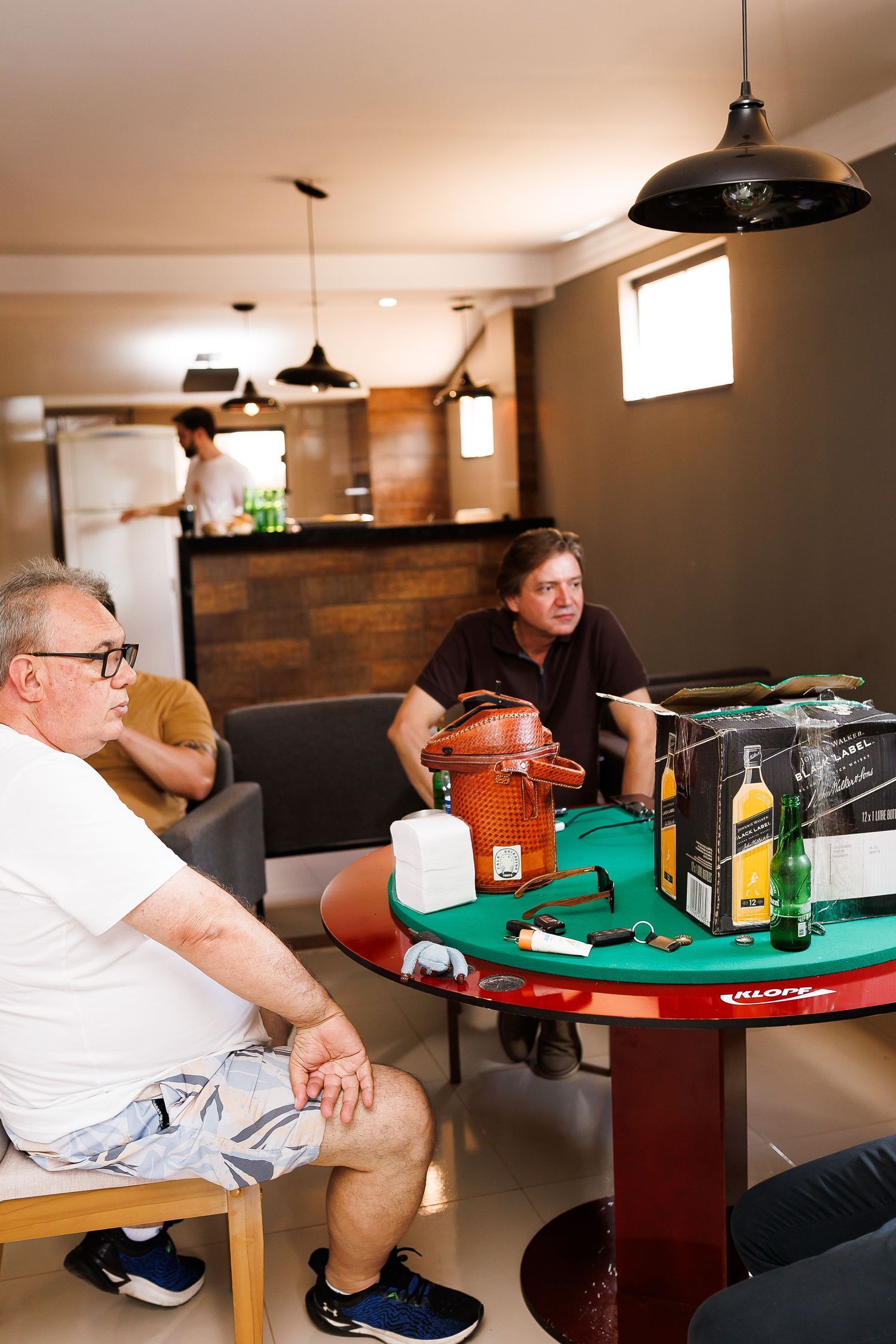 Two men sit at a green poker table with Johnnie Walker whiskey bottles in a modern lounge area.