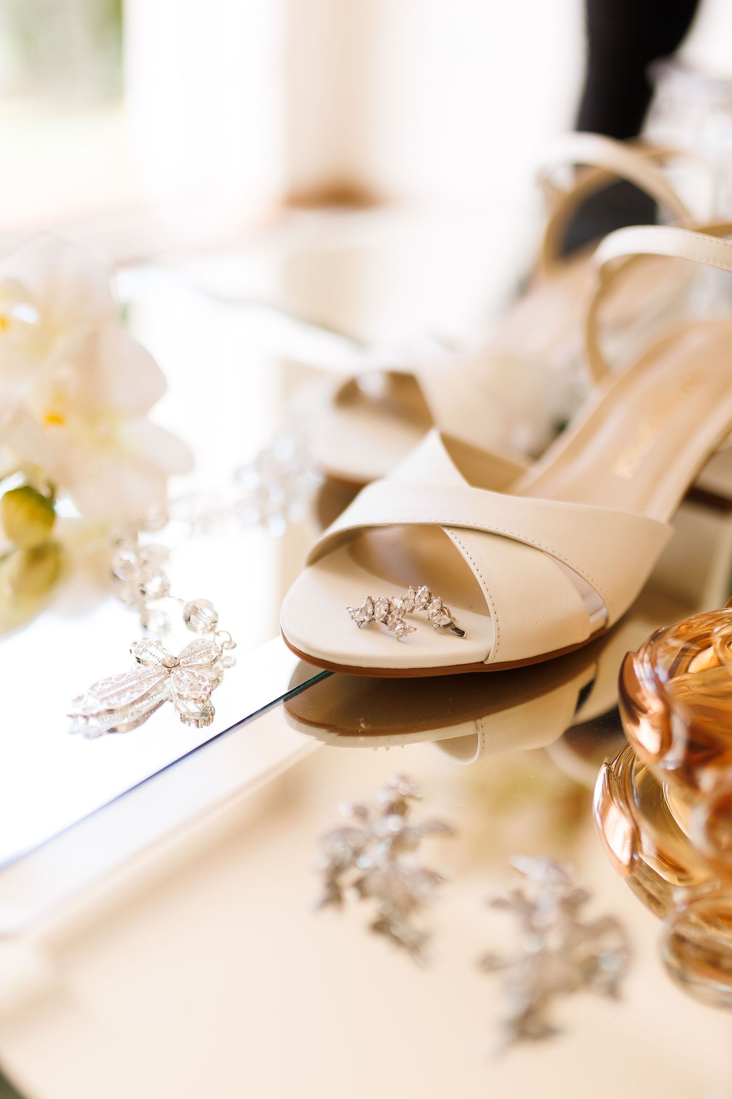 Elegant bridal heels with diamond ring and crystal jewelry on reflective surface with perfume and flowers.