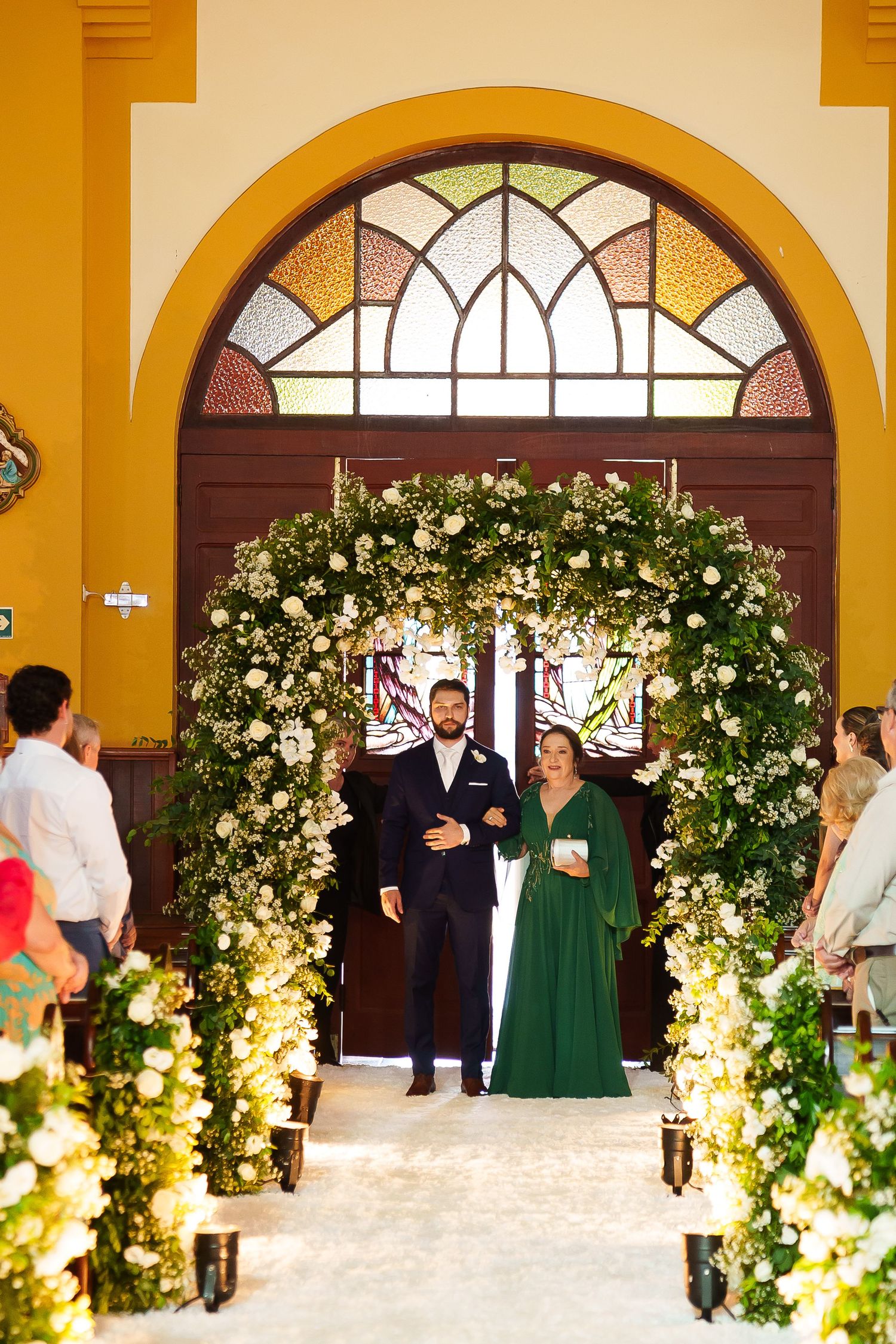 Groom and woman in green dress walk under floral arch at elegant church wedding ceremony with white flowers.