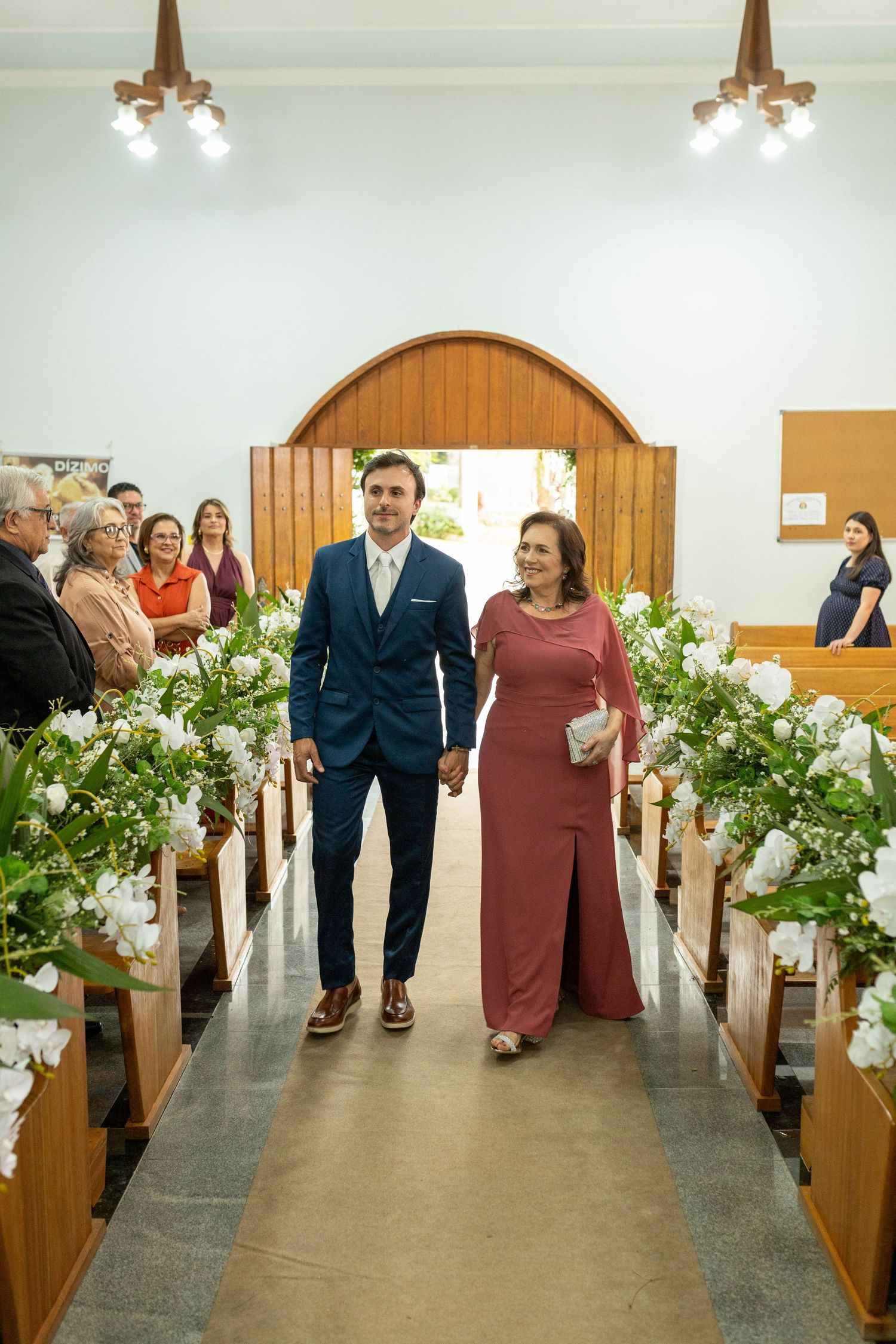 Groom in navy suit walks church aisle with woman in rose gown past white floral pew decorations.