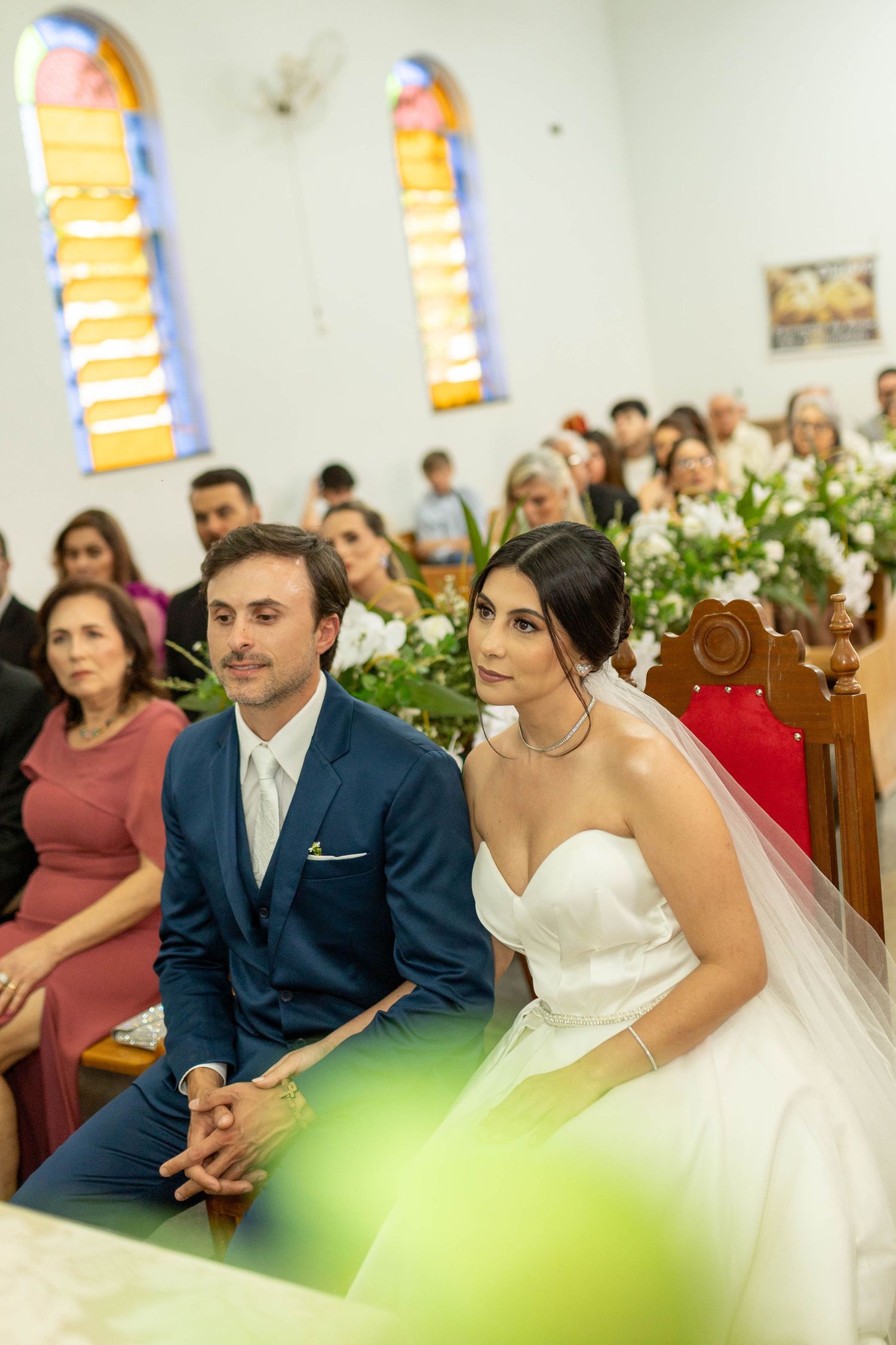 Bride in white gown and groom in navy suit sit together at church altar during wedding ceremony with guests seated behind.