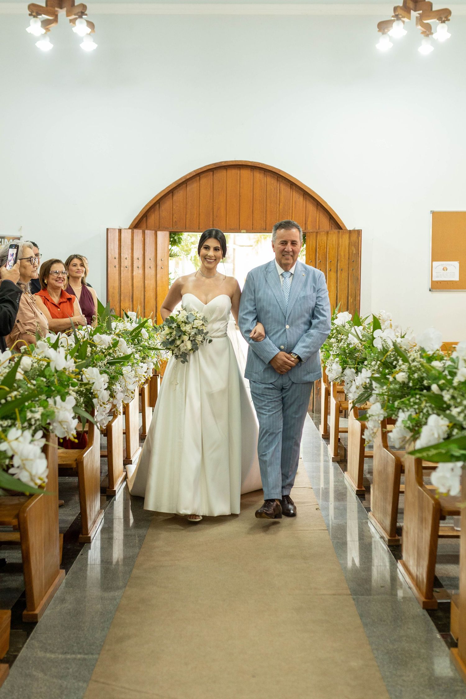 Bride in white gown walks down church aisle with escort, flanked by floral arrangements, during wedding ceremony.
