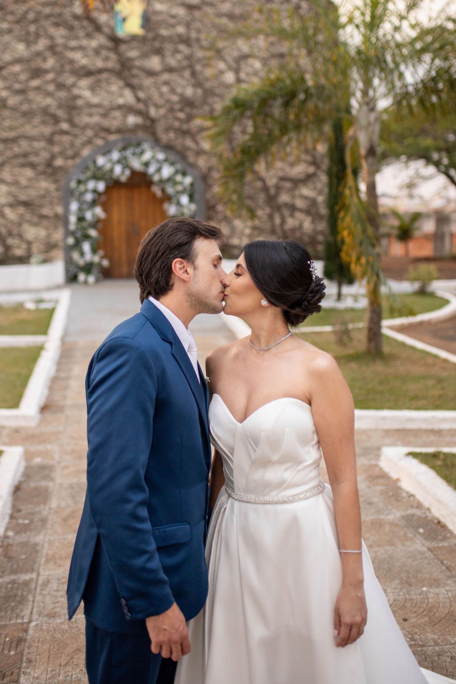 Bride and groom sharing a kiss outside a stone church, groom in blue suit, bride in strapless white wedding gown.