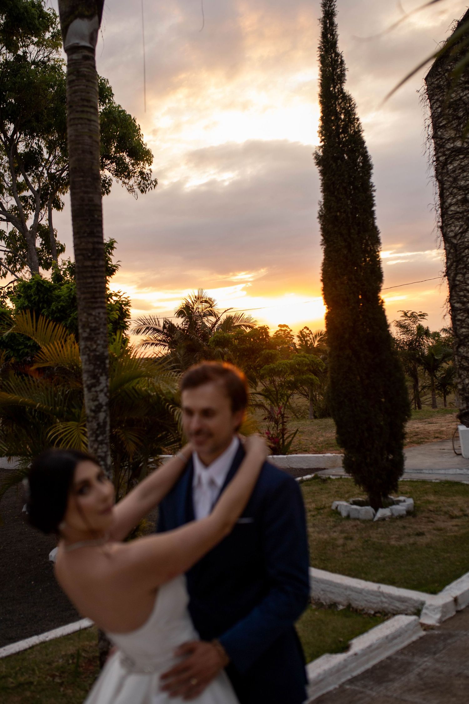 Bride and groom embracing at sunset in a lush garden with tall cypress trees and vibrant orange sky.