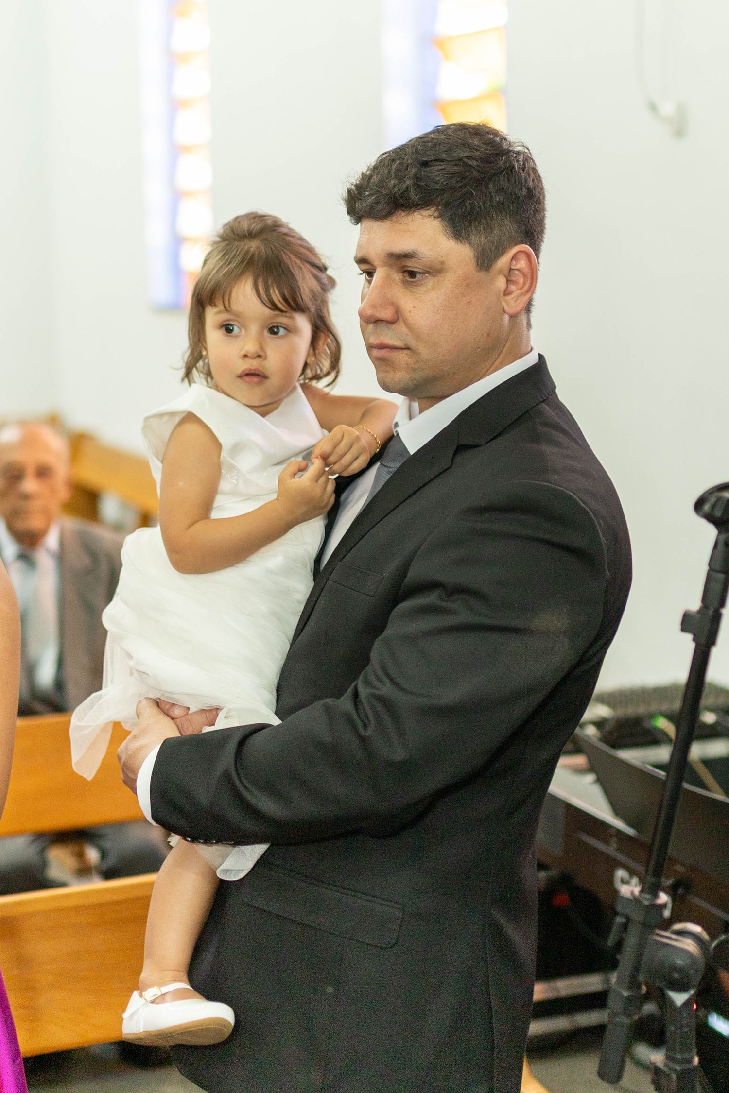 Man in dark suit holding a young girl in white dress inside a church during a formal ceremony.