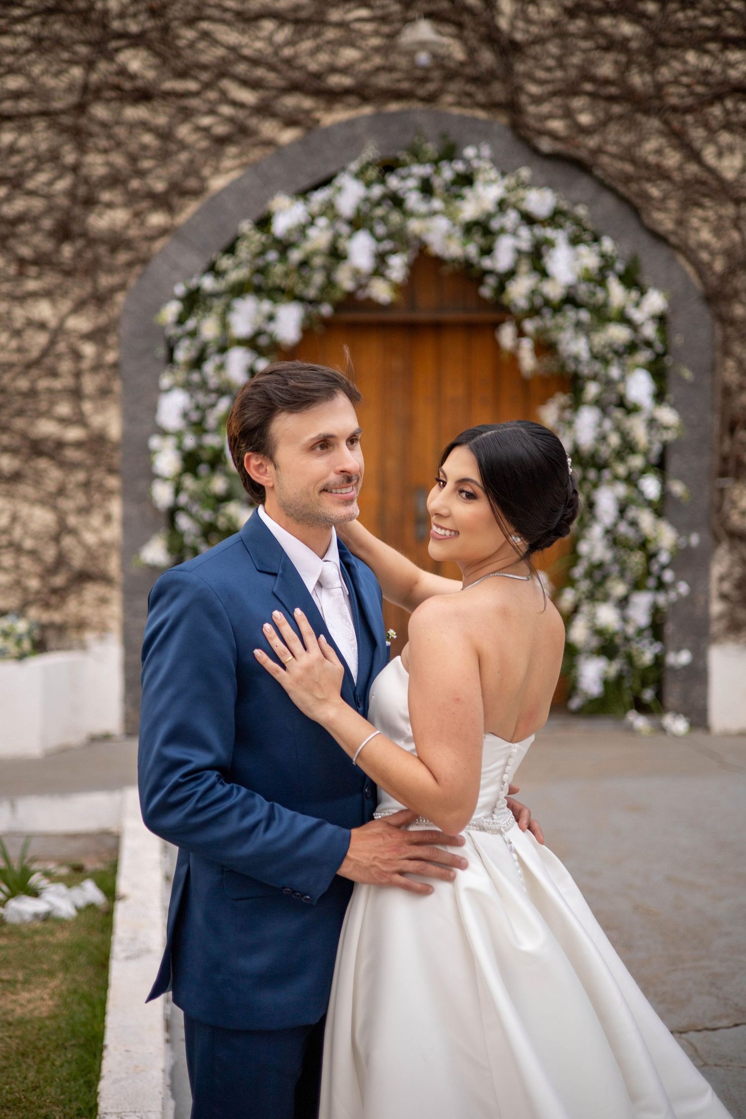 Bride and groom embrace in front of a floral arch at a wedding venue, she in a white gown, he in a blue suit.