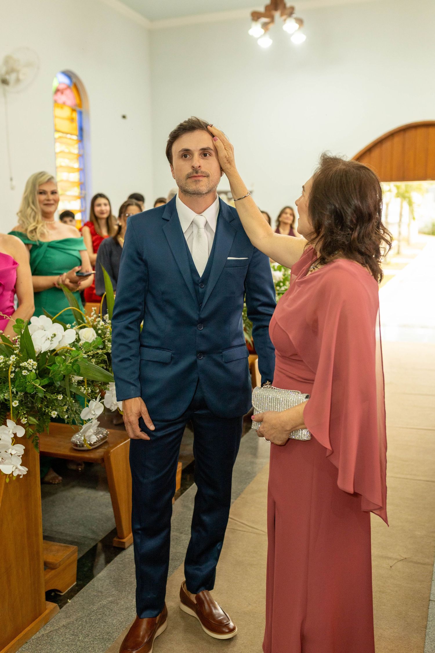Groom in navy suit receives a blessing from a woman in pink dress during a church wedding ceremony.