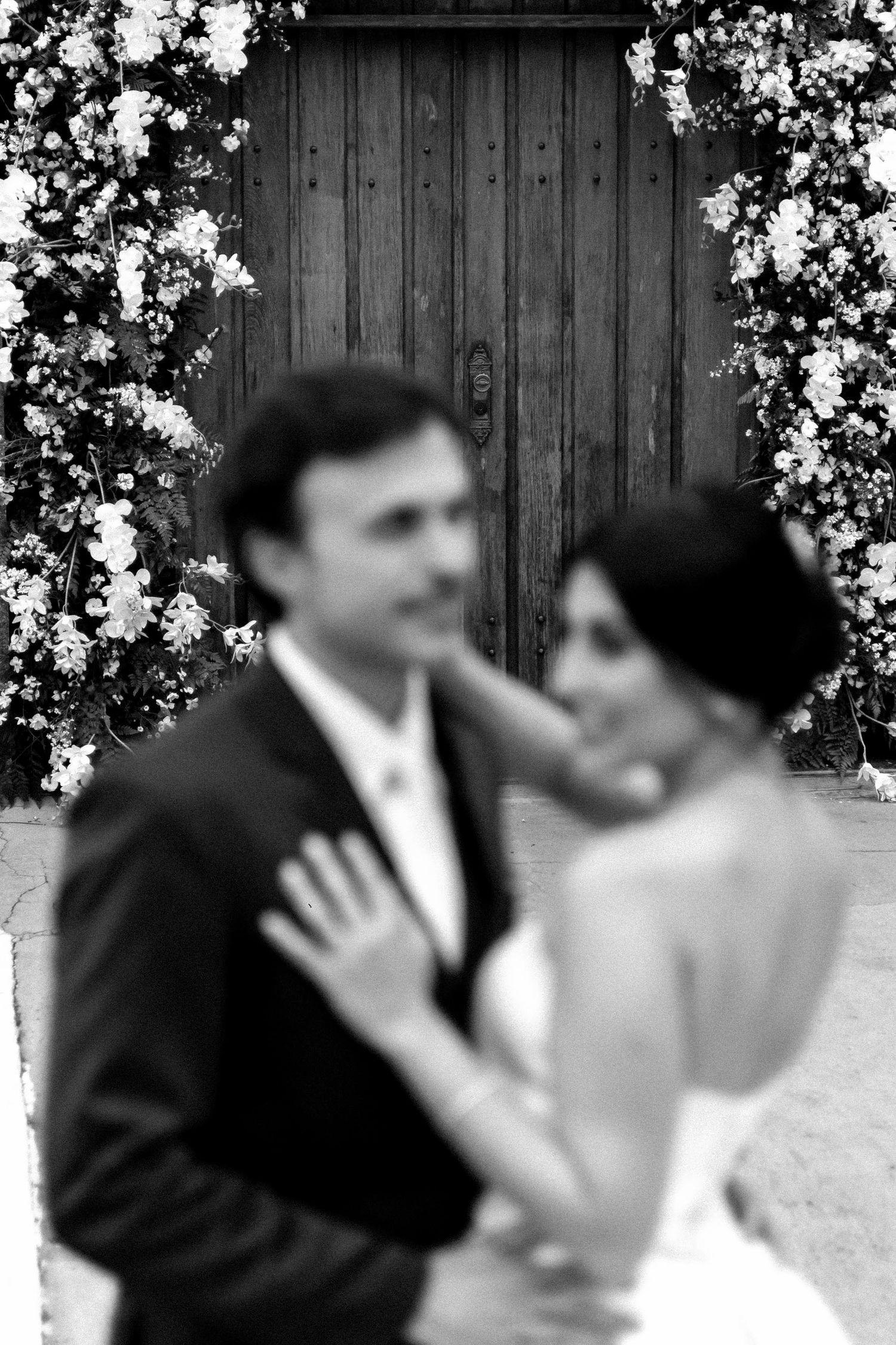 Black and white wedding photo of bride and groom embracing before a floral arch and rustic wooden door.