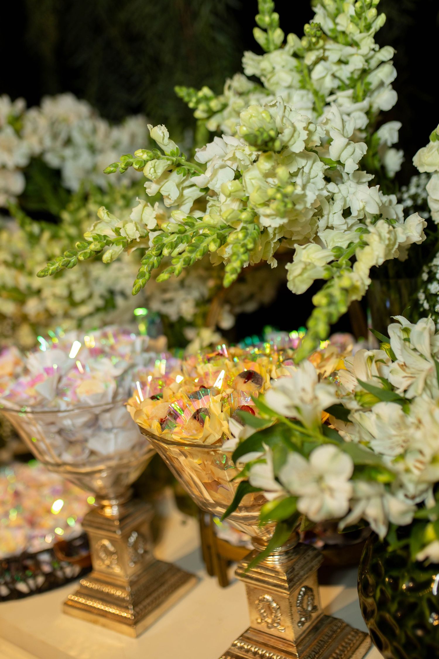 Elegant wedding candy buffet with gold pedestals holding sweets, surrounded by lush white snapdragon floral arrangements.