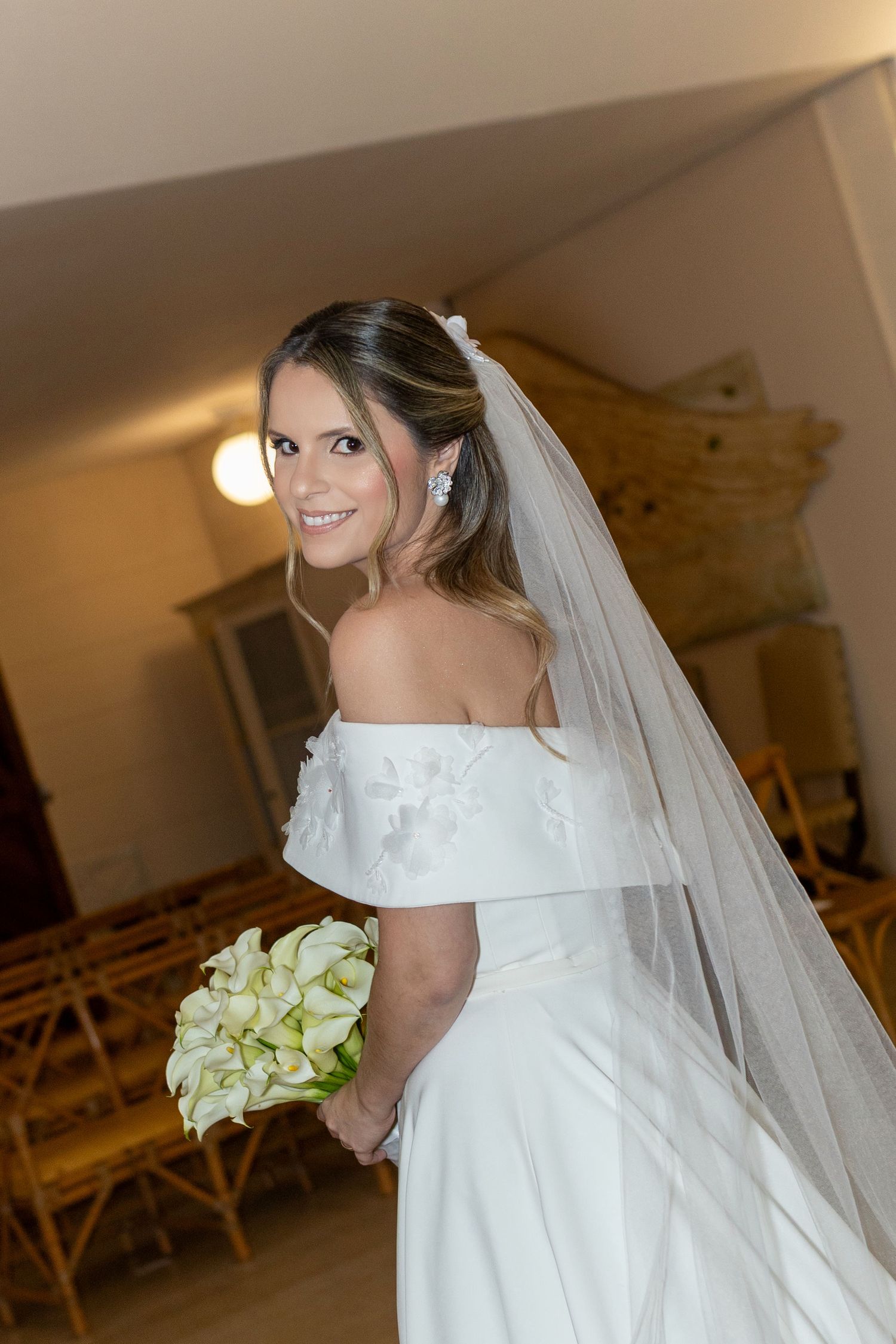 Smiling bride in off-shoulder white gown and veil holds calla lily bouquet, posing in a church aisle.