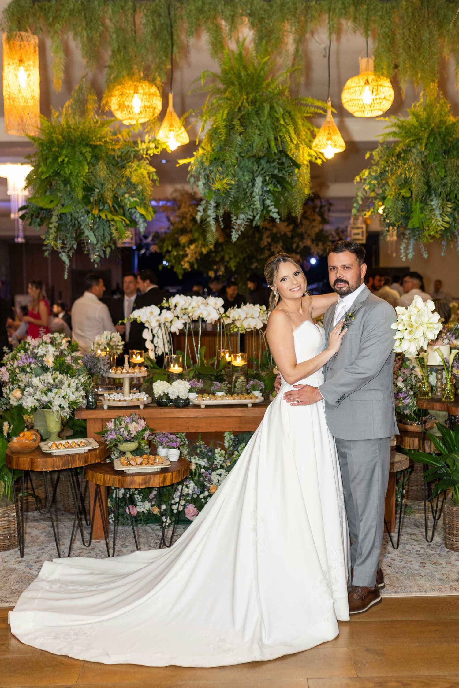 Bride and groom pose elegantly at their wedding reception surrounded by lush greenery, white orchids, and warm pendant lights.