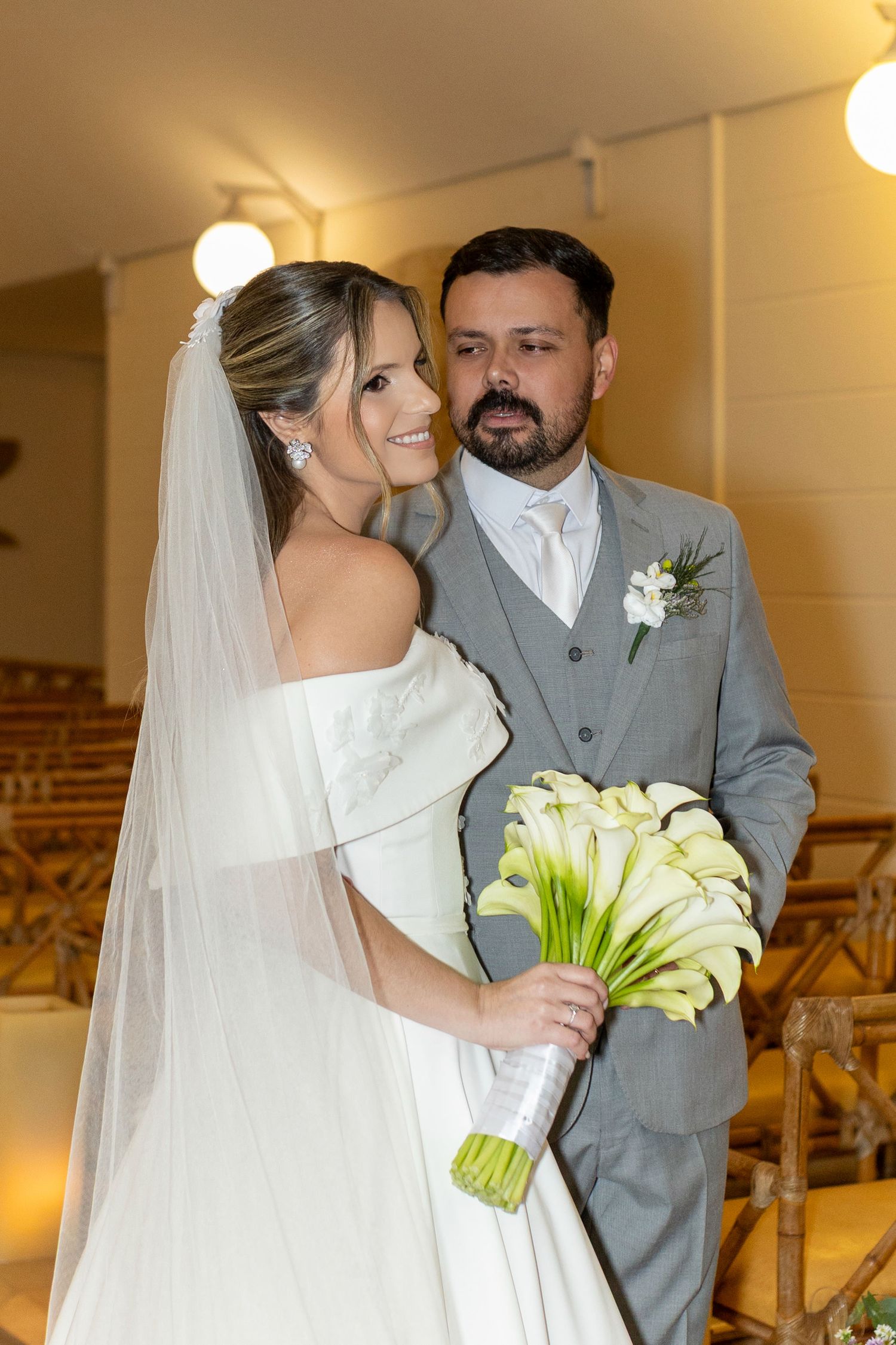 Bride in white off-shoulder gown with veil holds calla lily bouquet beside groom in gray suit at wedding ceremony.