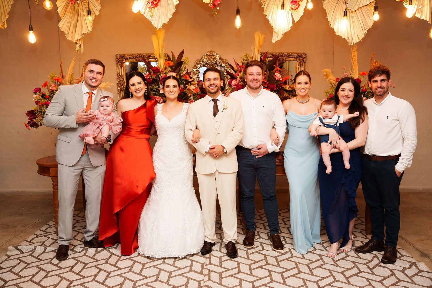 Wedding party group photo with bride in white gown, groom in tan suit, and guests in formal attire holding babies.