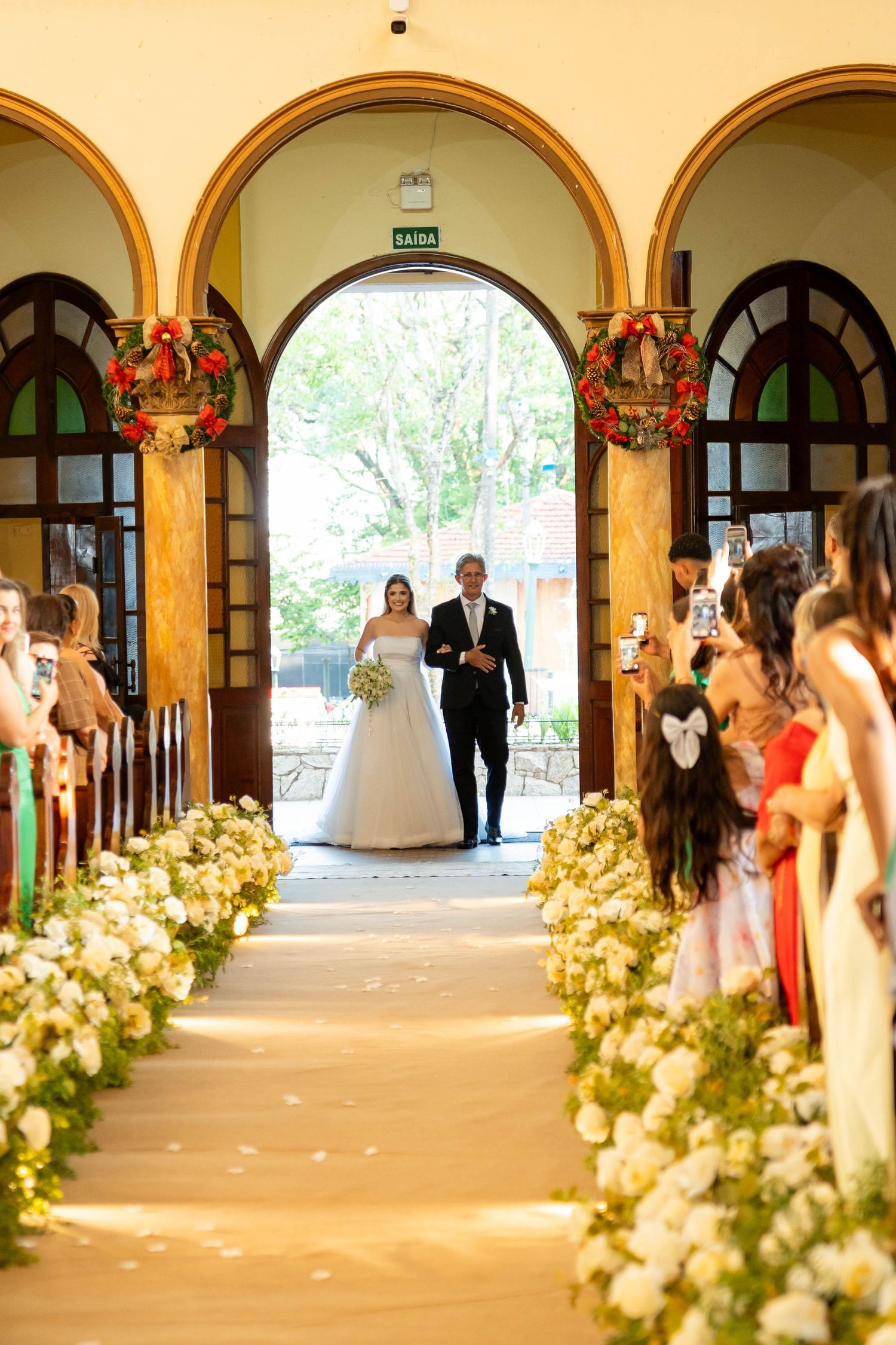 Bride walking down the aisle with escort in a beautiful chapel adorned with white flowers and arched doorways.