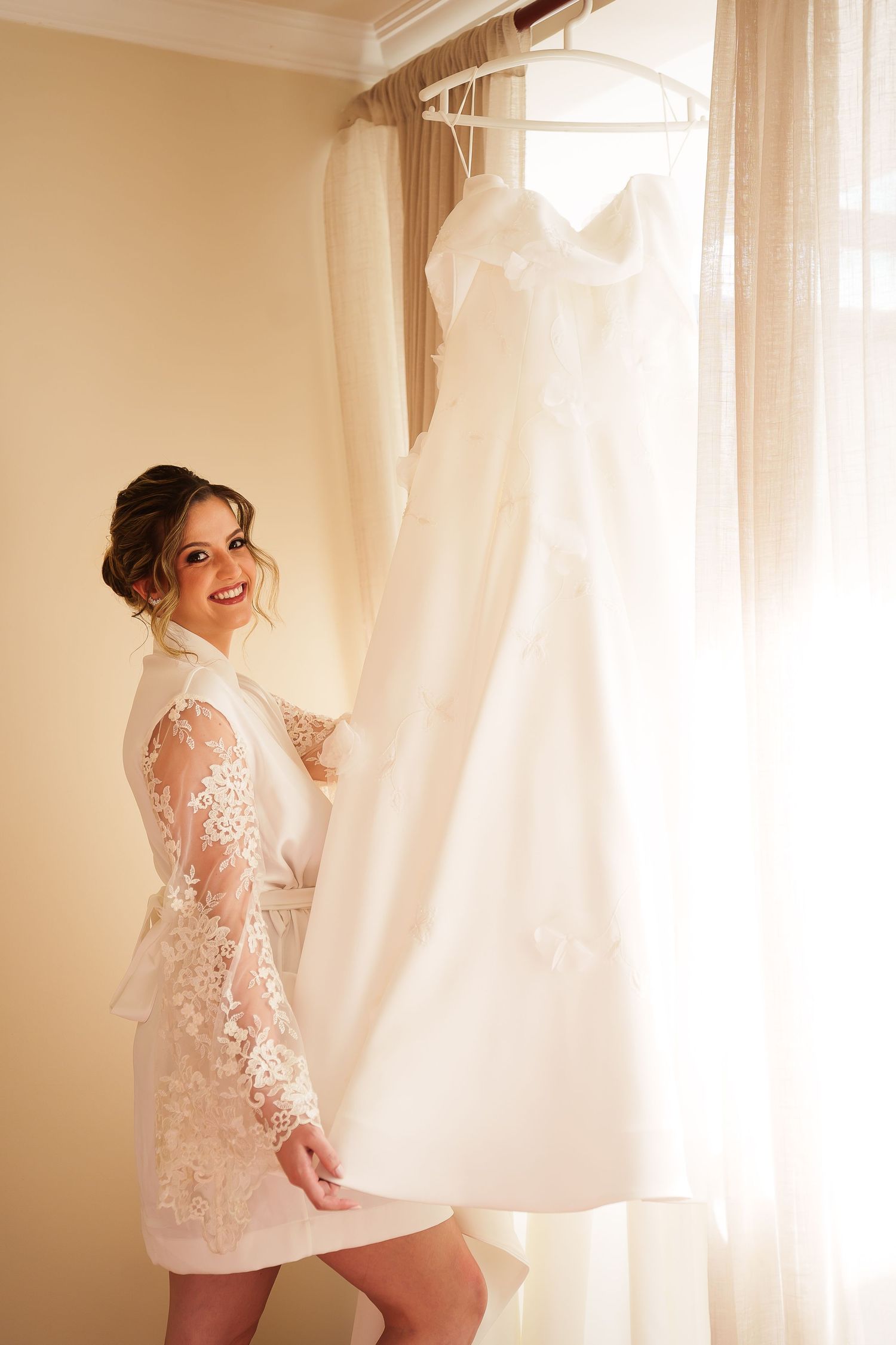 Smiling bride in lace robe standing next to her elegant white wedding dress hanging by a sunlit window.