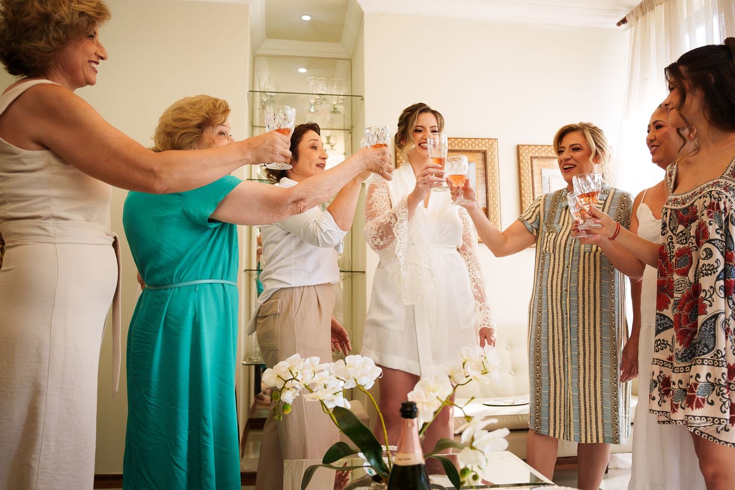 Bride in white robe toasting champagne with bridesmaids and family members during wedding preparation celebration indoors.