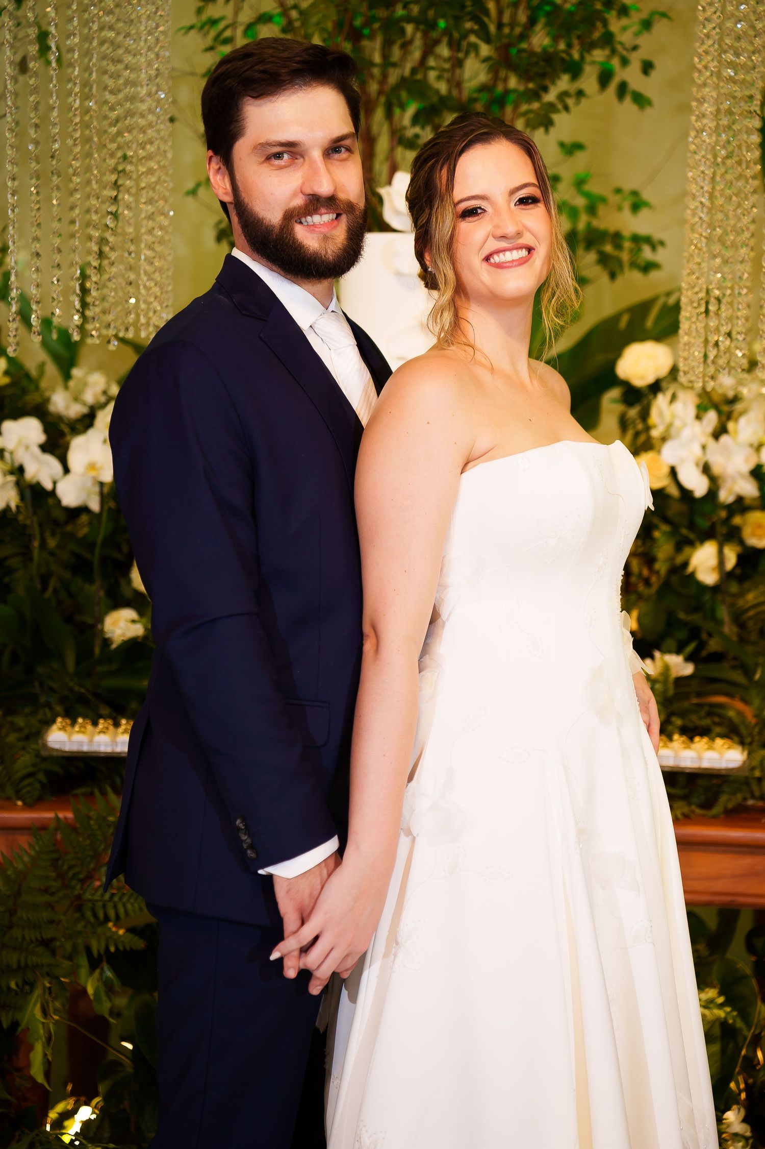 Smiling bride and groom pose together at their wedding reception, surrounded by lush greenery and white floral decor.