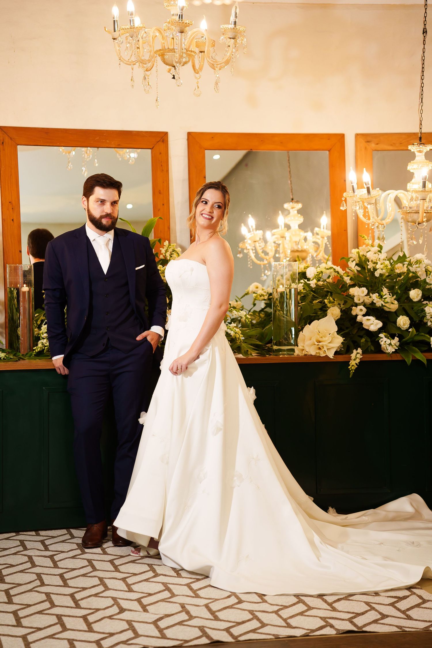 Bride in white gown and groom in navy suit pose elegantly in a chandelier-lit venue with floral arrangements.
