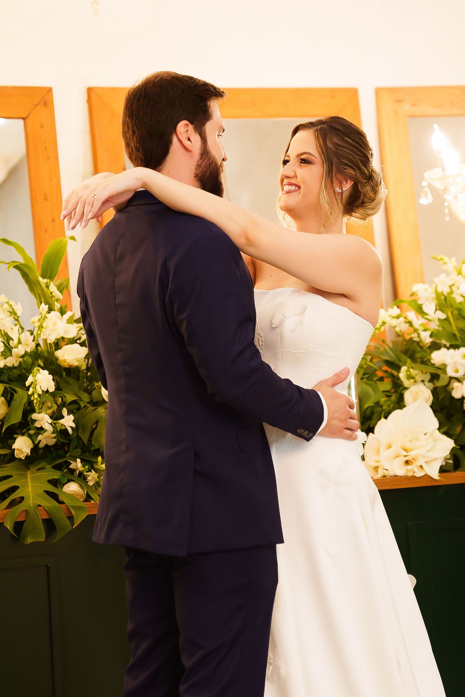 Bride and groom share a romantic embrace, smiling at each other, surrounded by white floral arrangements.