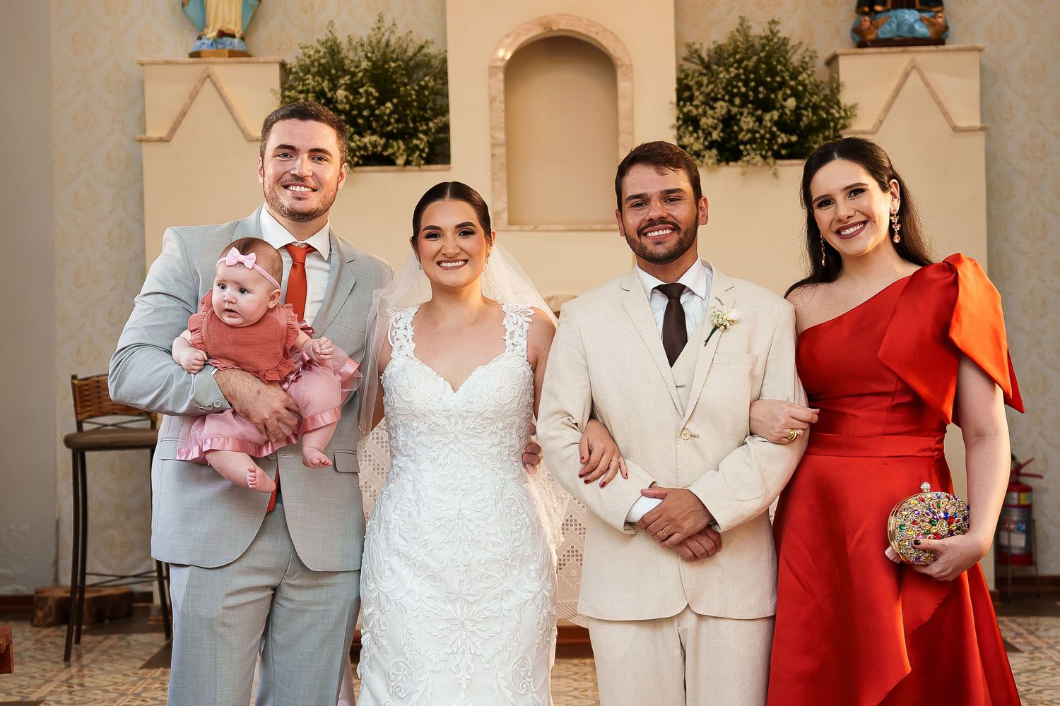 Wedding party poses in church, bride in lace gown, groom in cream suit, guests in gray suit and red dress holding baby.