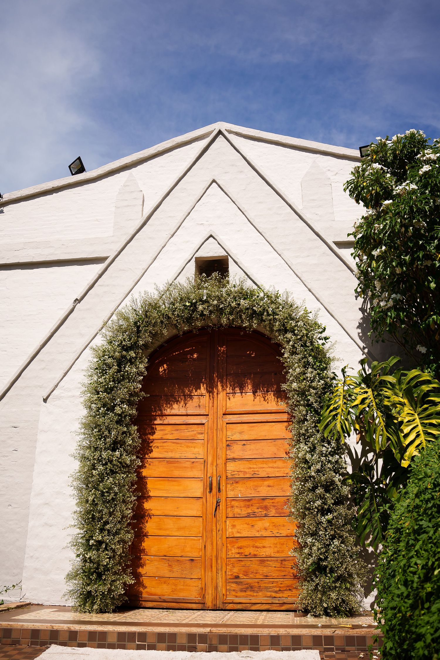 White church building with arched wooden doors framed by lush green foliage against a blue sky.