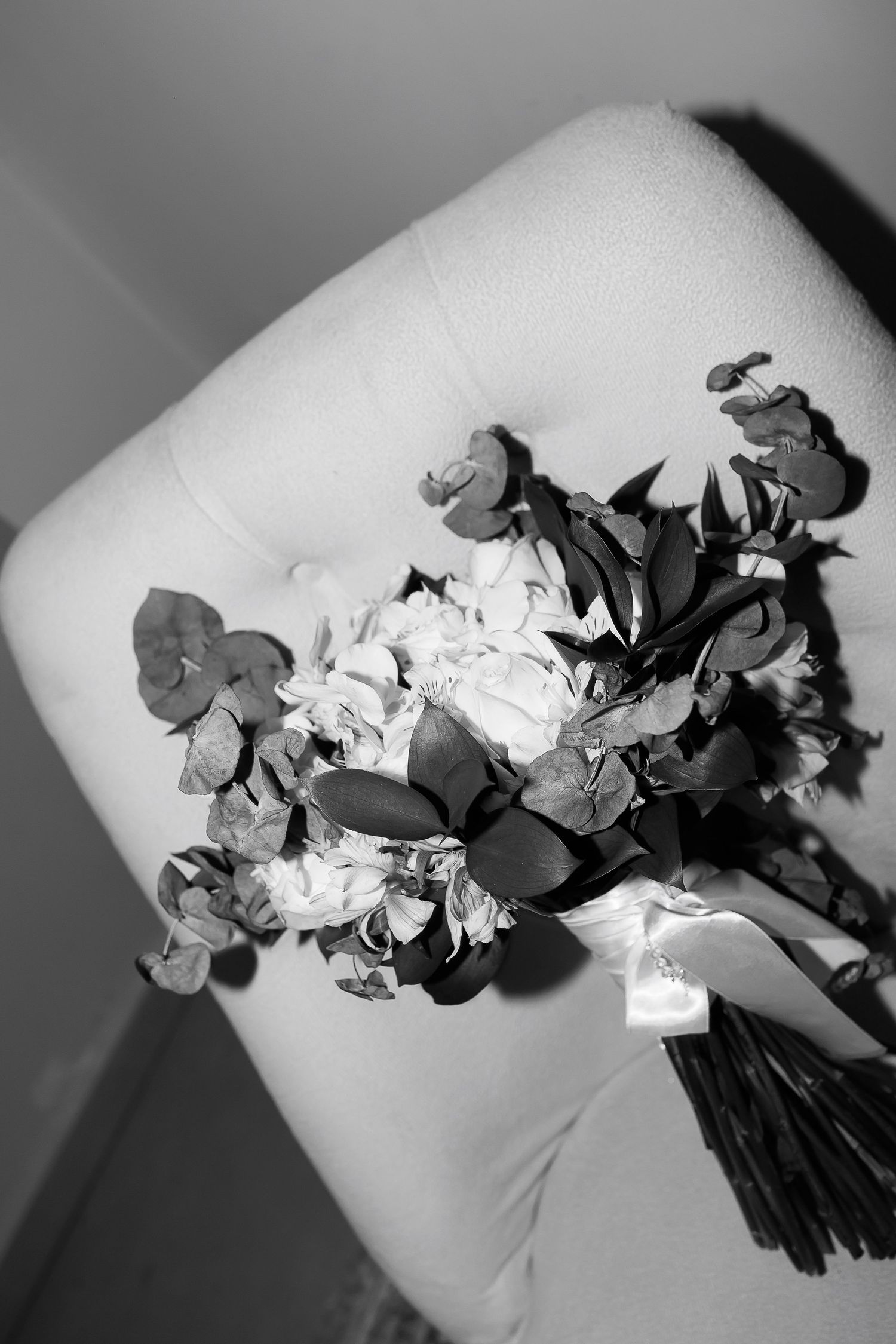 Black and white photo of a bridal bouquet with roses and eucalyptus resting on a white upholstered chair.