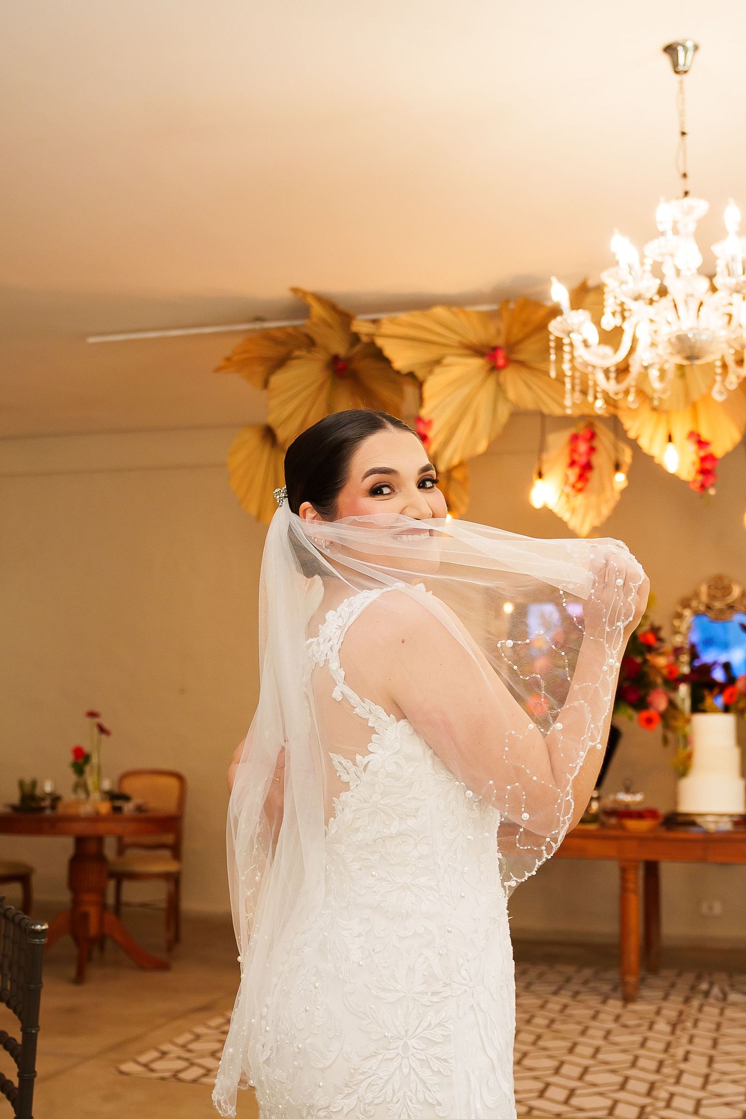 Bride in lace wedding dress holding veil over face, posing in elegant venue with chandelier and floral decor.