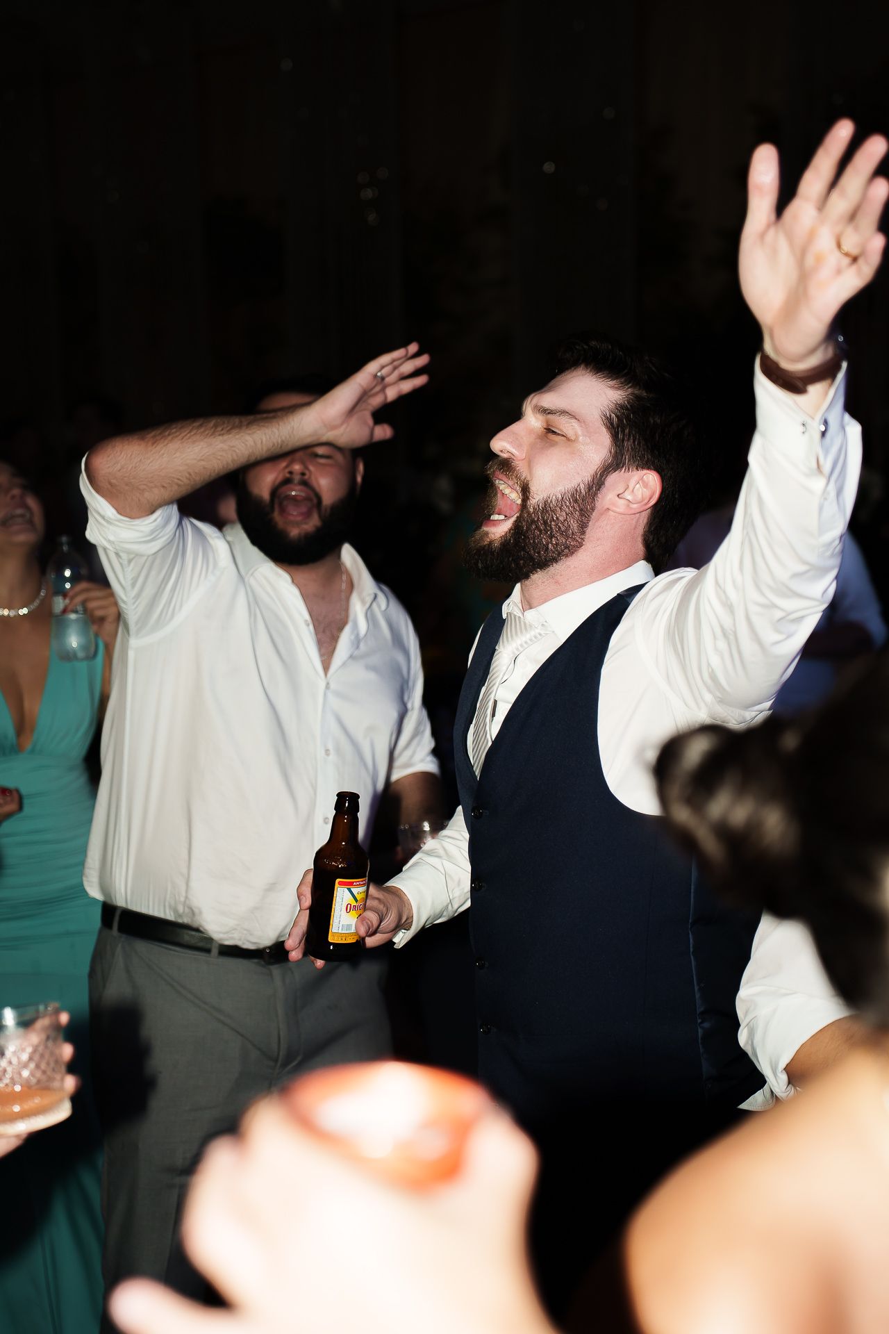 Men dancing enthusiastically at a wedding reception, one holding a beer bottle with arms raised in celebration.