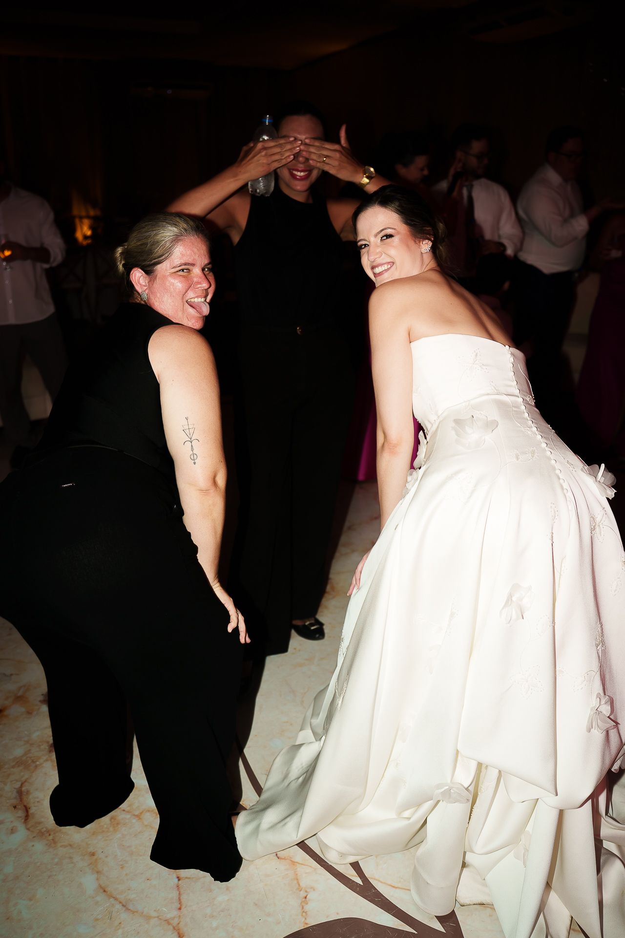 Bride in white gown laughs with guests at wedding reception while man playfully covers his eyes.