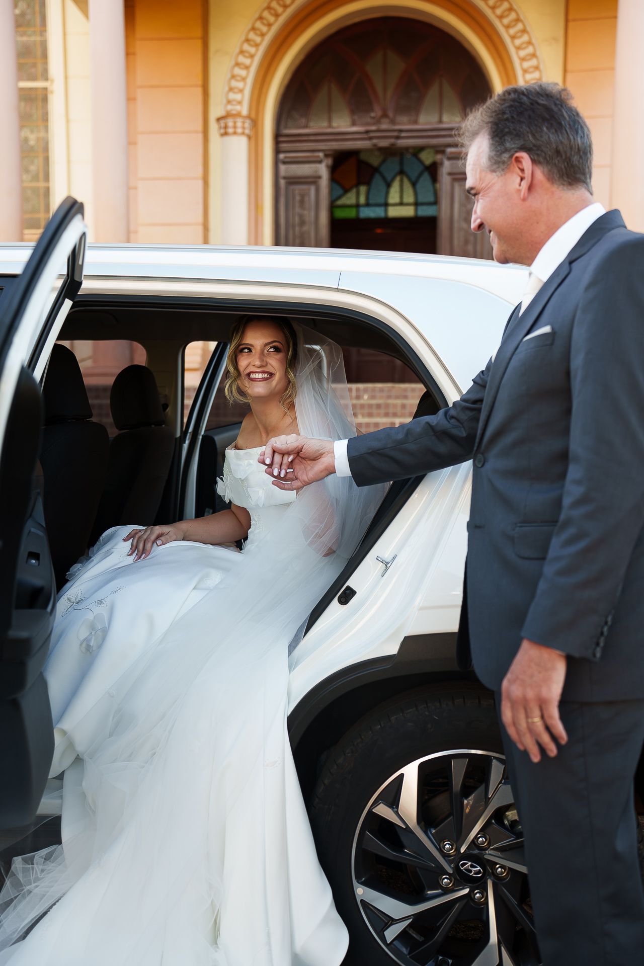 A smiling bride in a white gown exits a limousine, assisted by a man in a suit, outside a church.