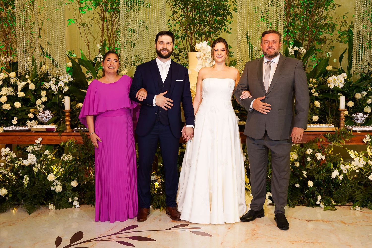 Elegant wedding party of four posing together, bride in white gown, groom in navy suit, guests in pink and gray formal attire.
