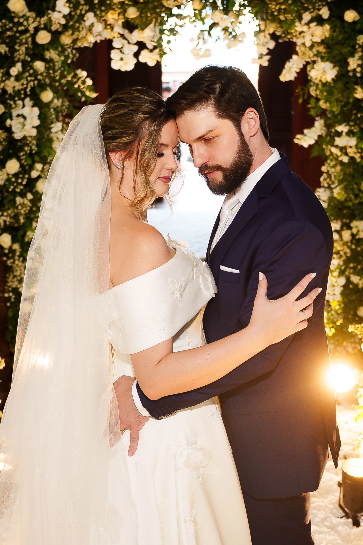 A bride and groom share a romantic embrace at their wedding ceremony beneath a floral arch with warm golden lighting.