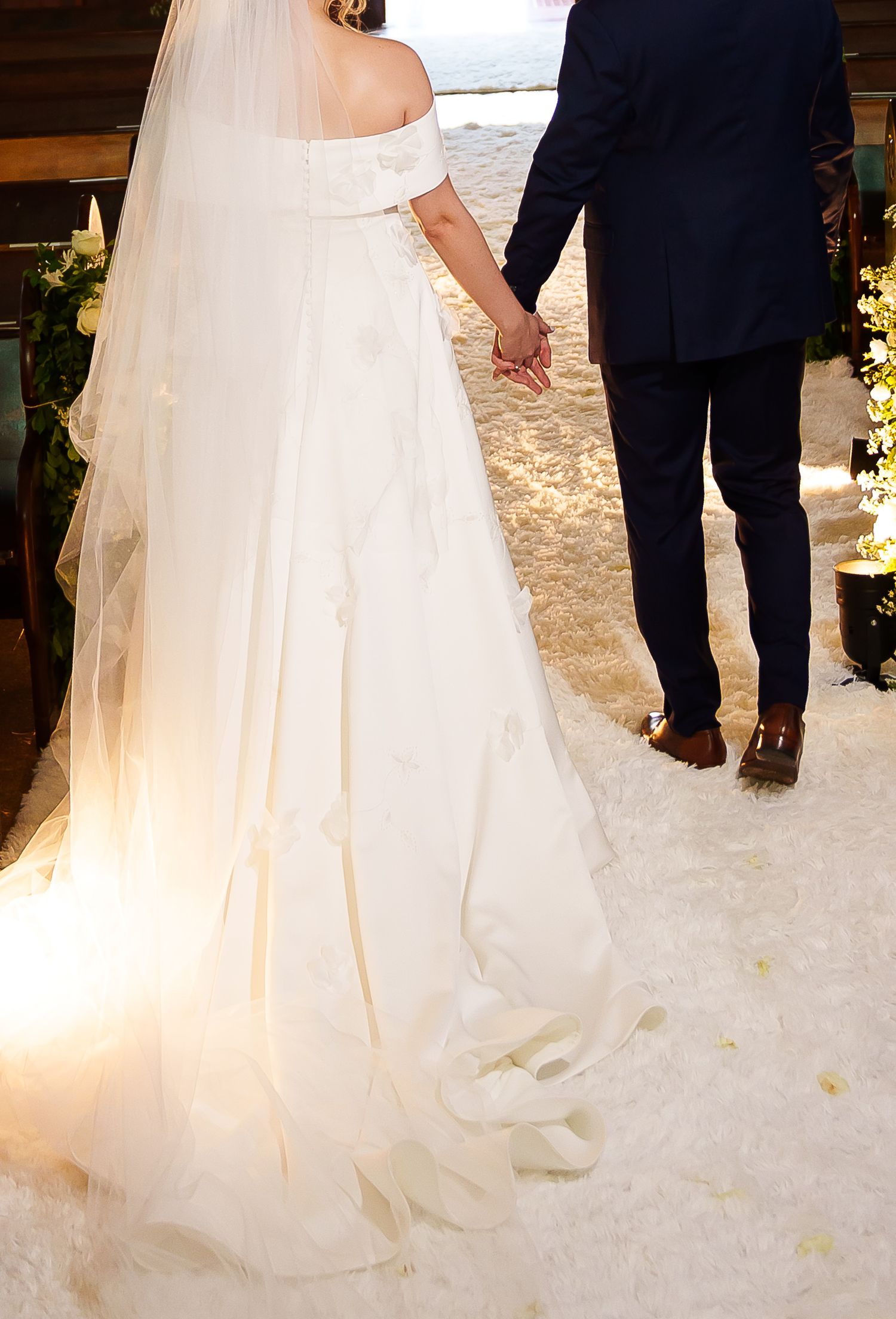 Bride in off-shoulder white gown with veil holds groom's hand in navy suit at elegant wedding ceremony.