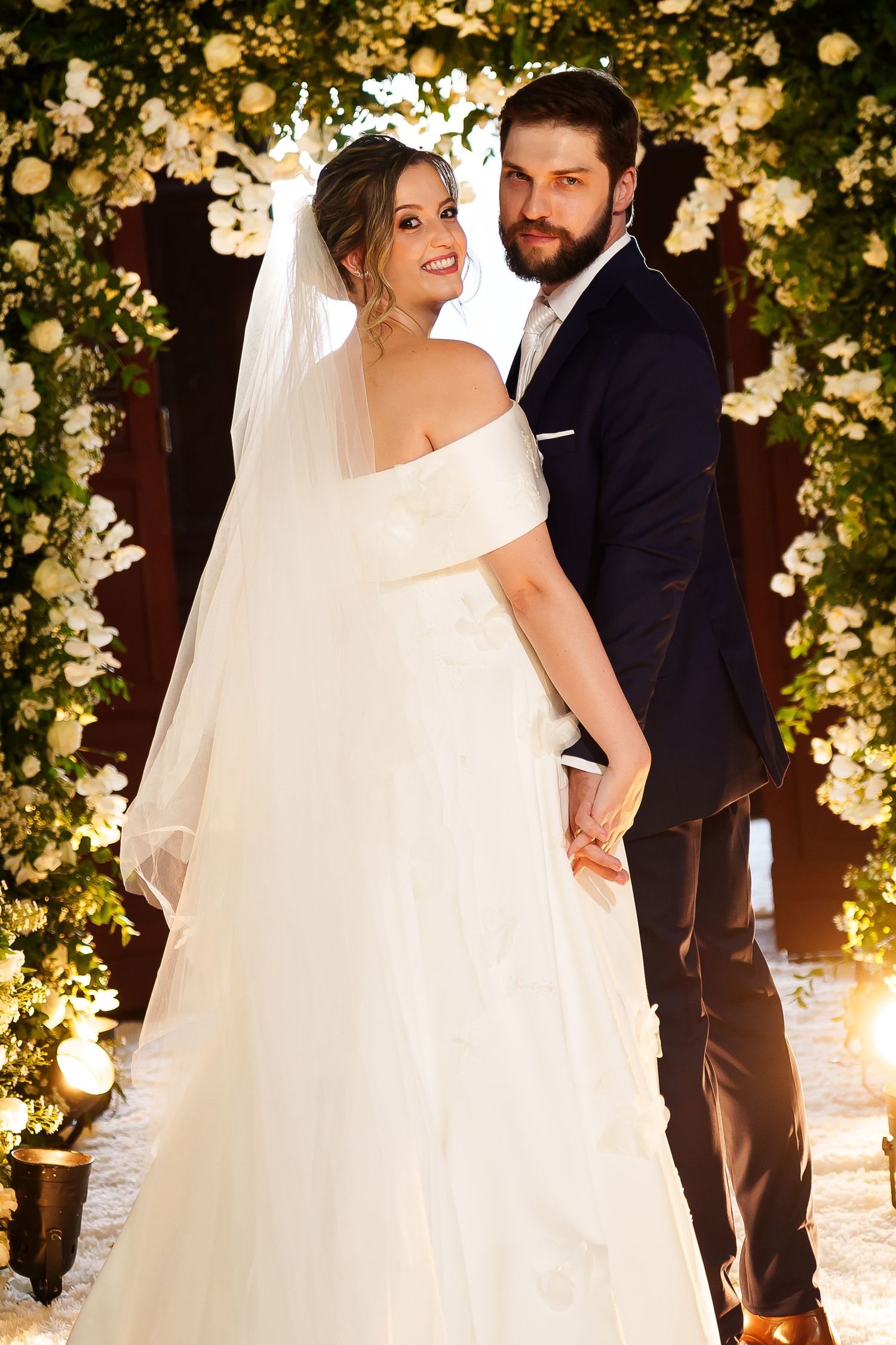 Bride in white off-shoulder gown and veil poses with bearded groom in navy suit before floral arch.
