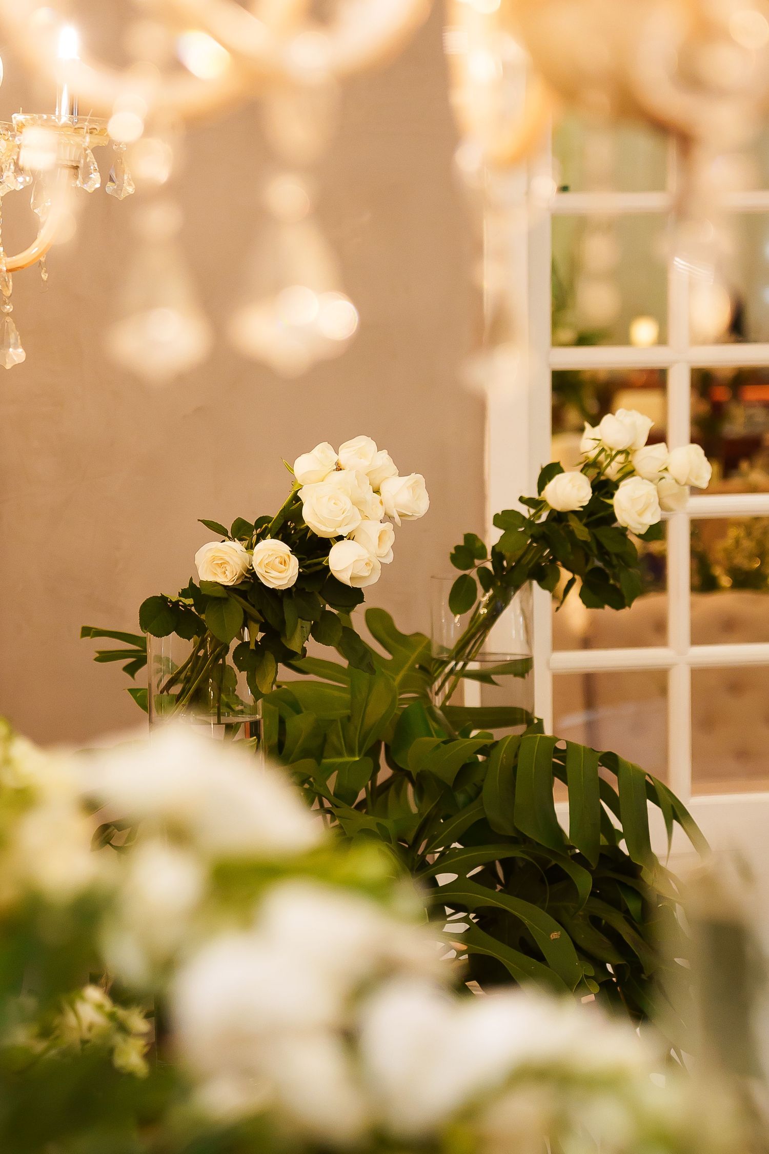 Elegant white roses with green leaves arranged near a window, with a crystal chandelier casting warm light.